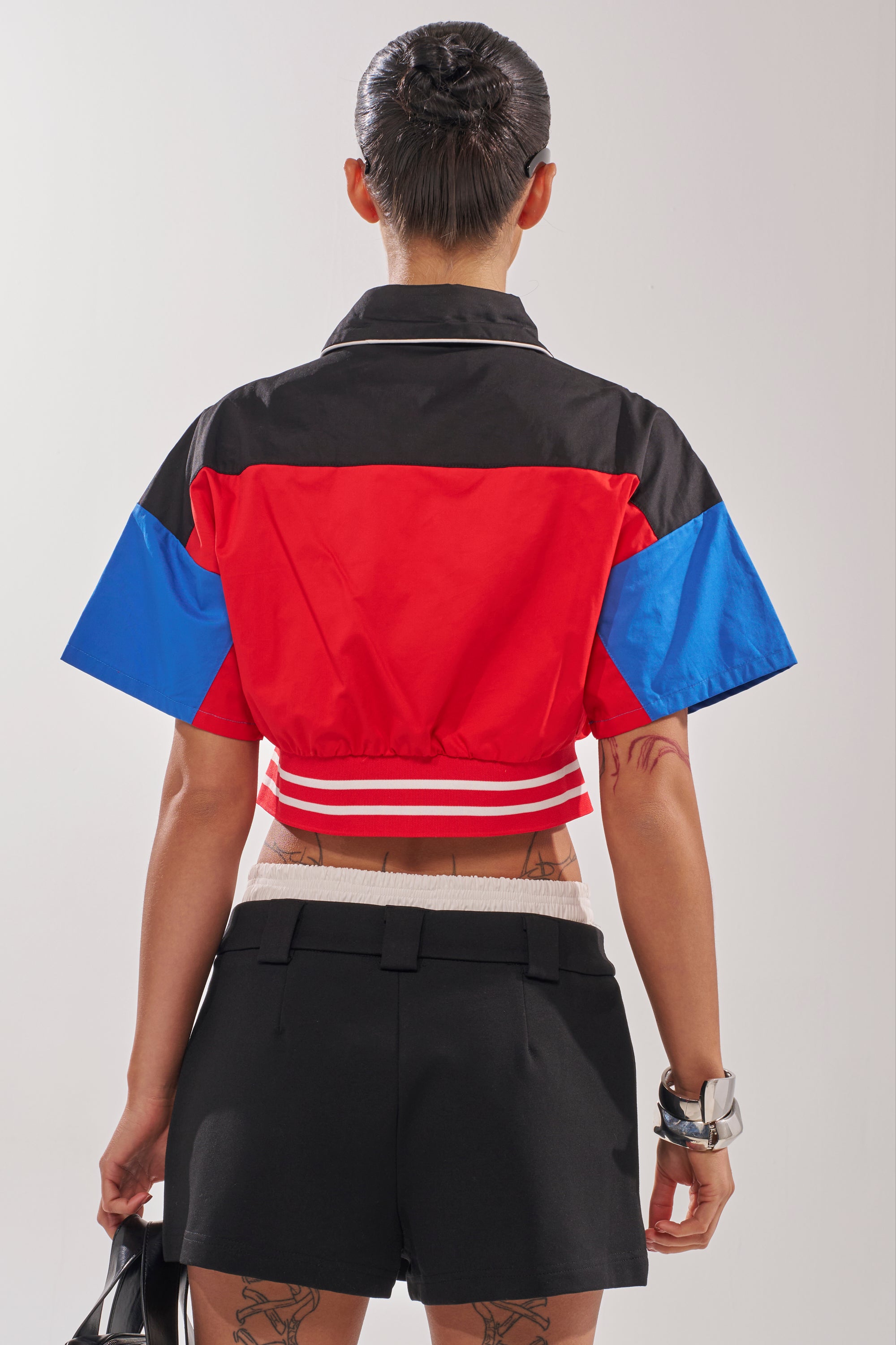A person with short hair is seen from behind wearing the HIGH GEAR CROPPED BUTTON DOWN in red, black, and blue, paired with black high-waisted shorts. Tattoos are visible on their legs, and they sport a chunky silver bracelet. 4