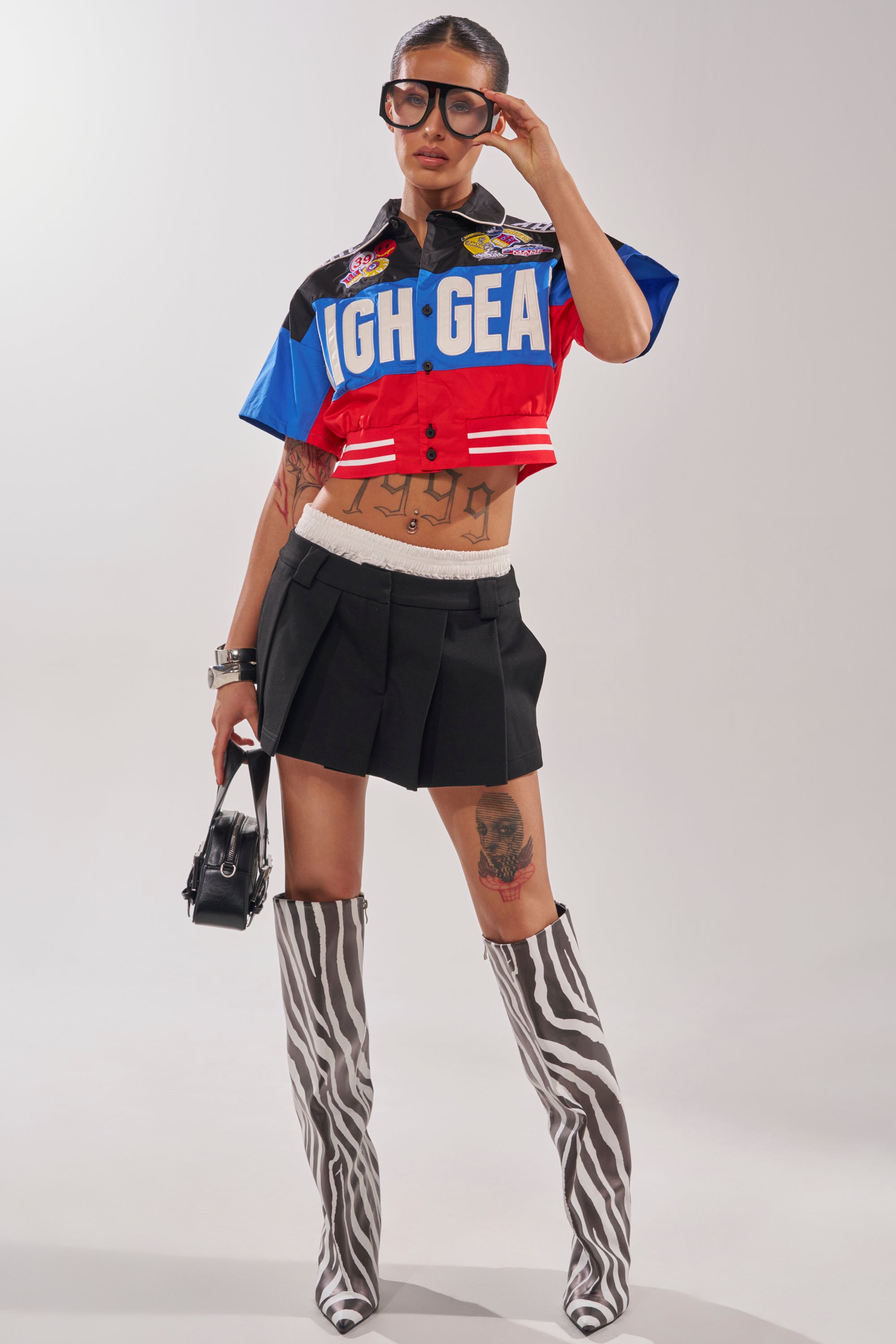 Wearing oversized glasses, a HIGH GEAR CROPPED BUTTON DOWN in vibrant streetwear style, a black pleated mini skirt, zebra-print knee-high boots, and a small black handbag, someone poses confidently against a plain background. 5