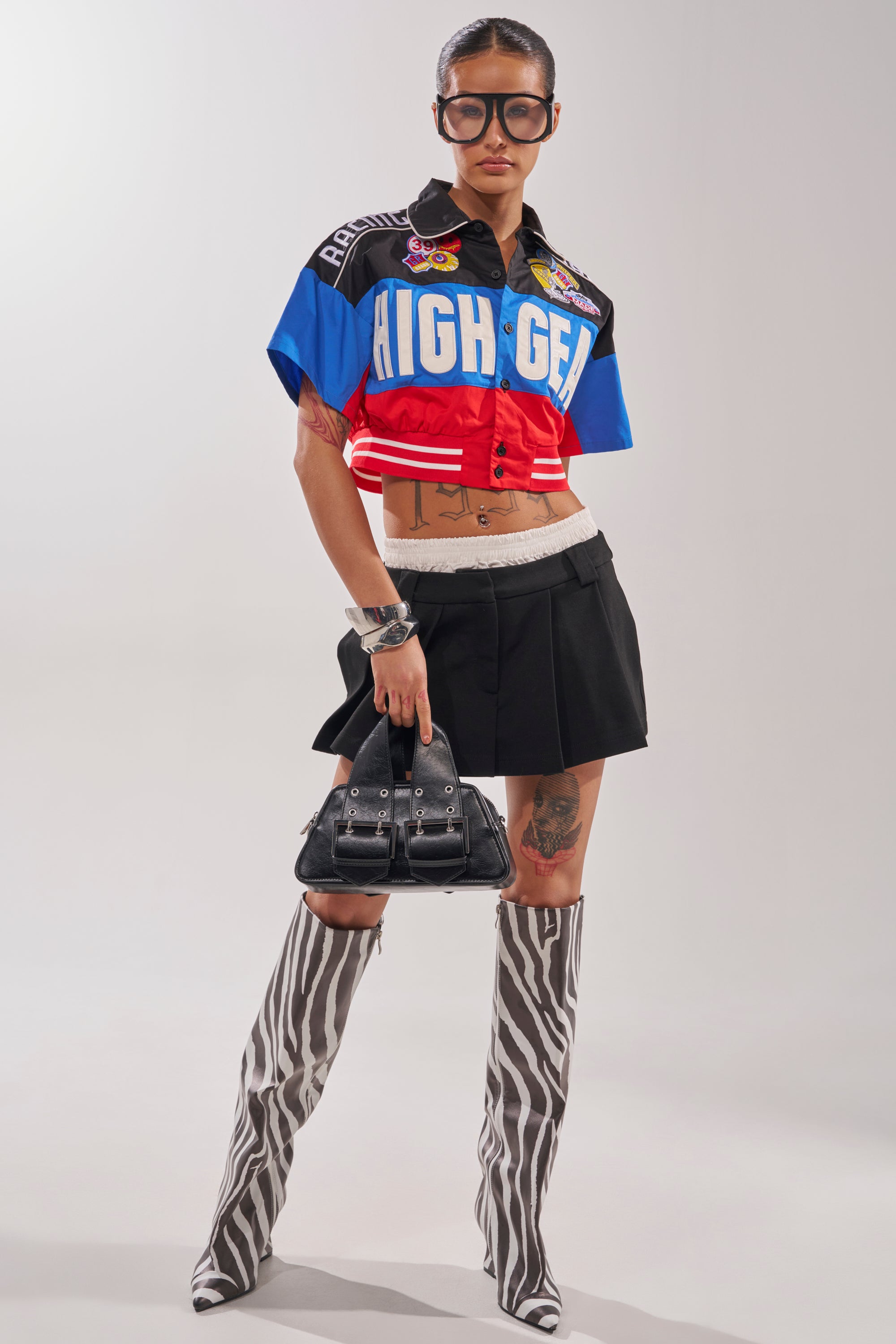 A woman in oversized glasses, the HIGH GEAR CROPPED BUTTON DOWN jacket with "HIGH GEAR" text, a black mini skirt, zebra-print knee-high boots, and a black handbag stands confidently against a plain backdrop. 6