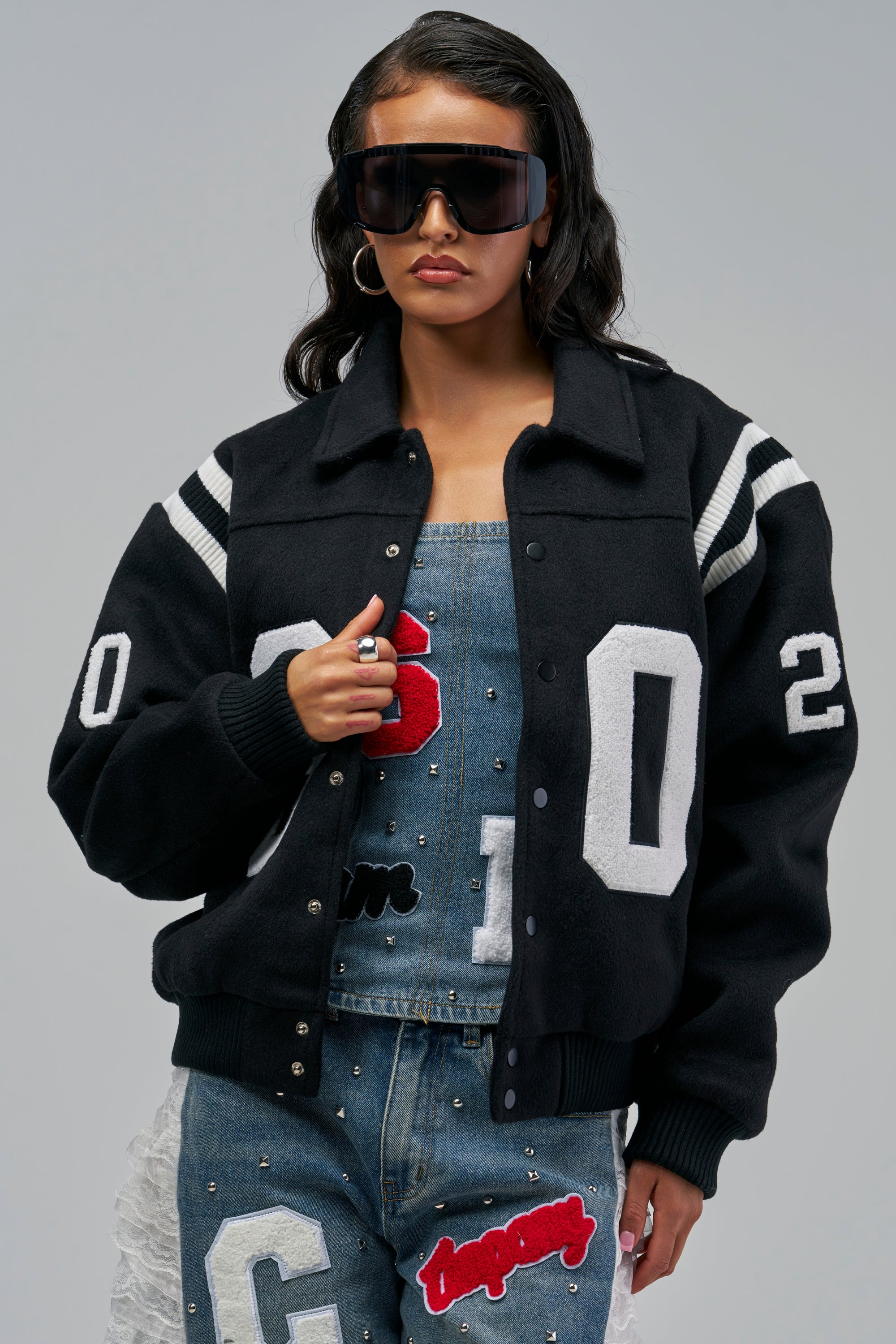 A person wearing a black CLUB LEVEL VARSITY BOMBER jacket with white striped cuffs and white numbers on the sleeves and chest.