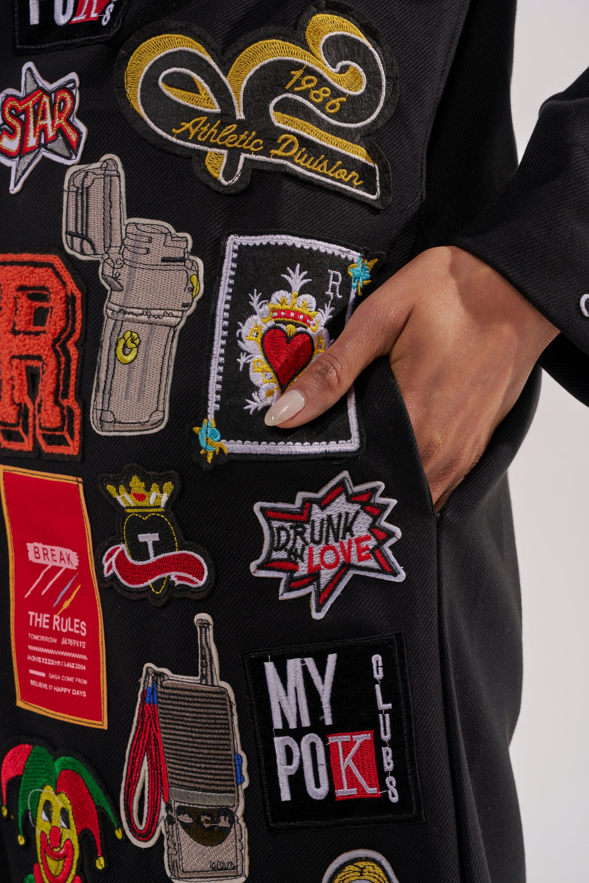 A close-up of a manicured hand rests in the pocket of the EVERYWHERE I AM PATCHWORK TRENCH, a black coat featuring colorful embroidered patches, bold phrases, and playful designs—true statement outerwear. 9