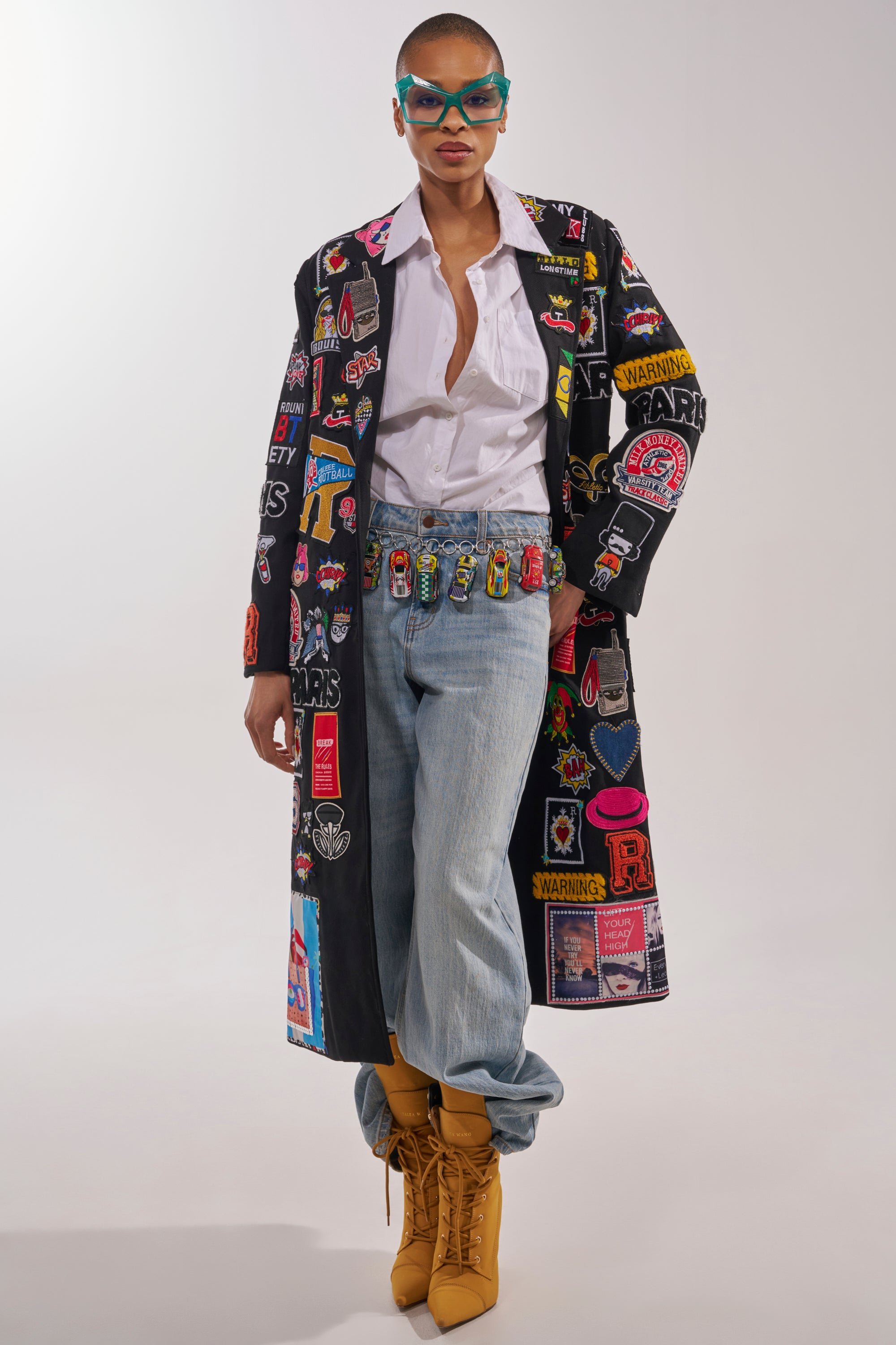 Wearing the EVERYWHERE I AM PATCHWORK TRENCH, a white shirt, blue jeans, tan lace-up boots, blue sunglasses, and a doll-embellished belt, a person stands confidently against a neutral background. 1