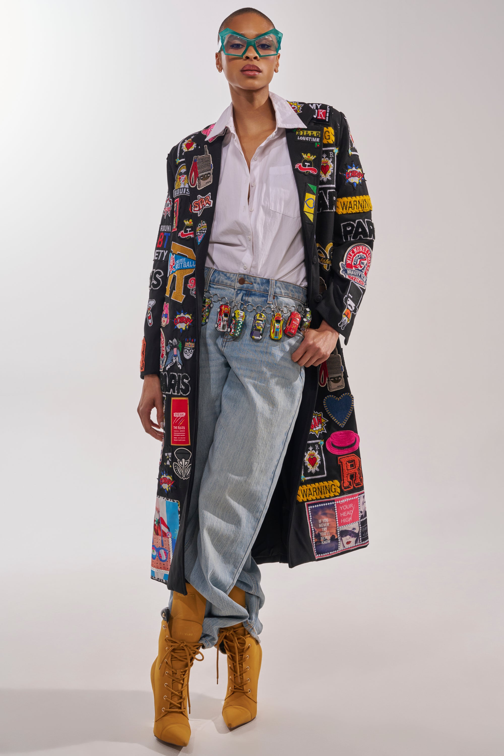 A stylish person poses in the EVERYWHERE I AM PATCHWORK TRENCH, light blue jeans, a white shirt, tan lace-up boots, and blue glasses against a plain background—a true nod to maximalist street style. 2