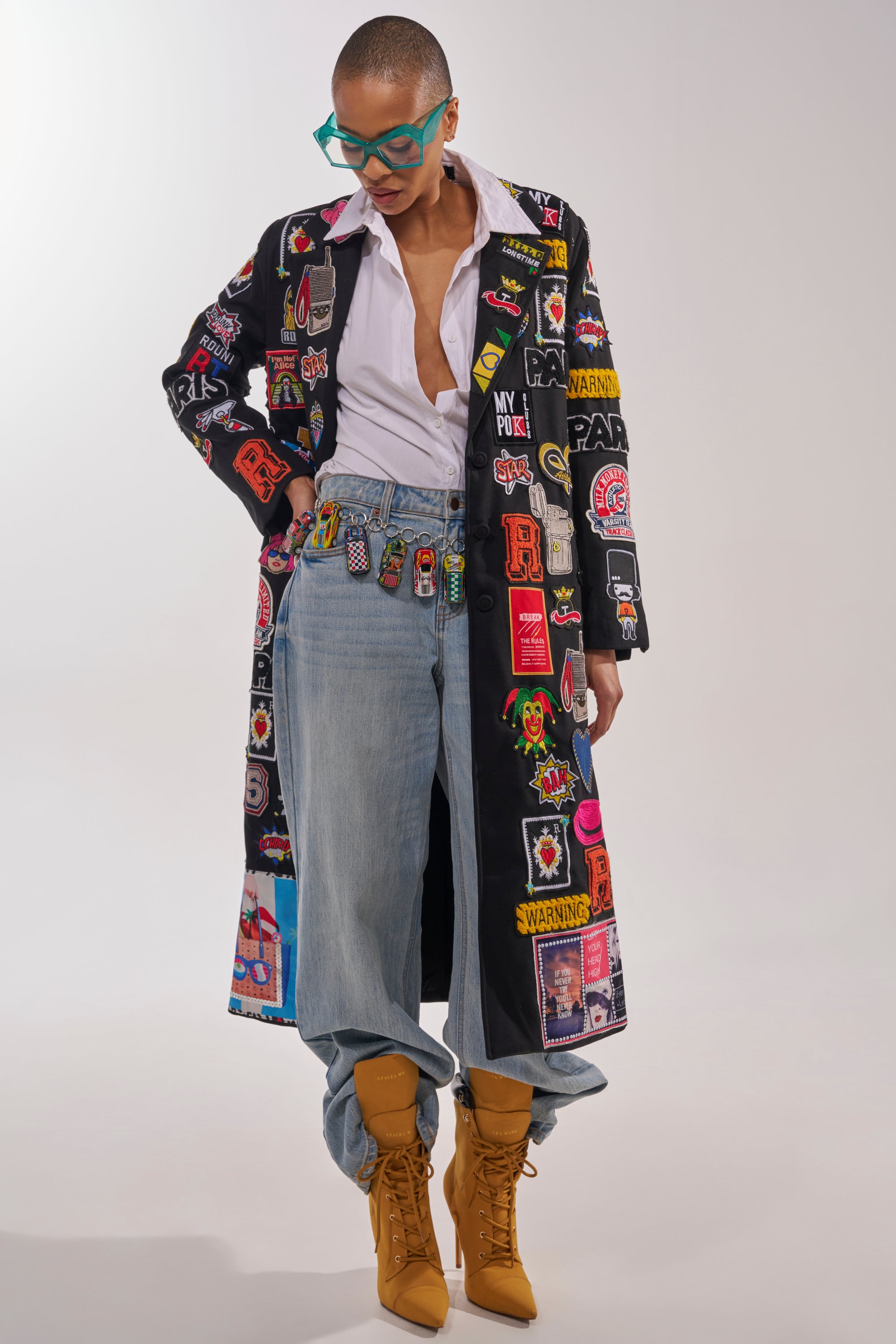 A person with short hair and teal glasses wears the EVERYWHERE I AM PATCHWORK TRENCH over a partly unbuttoned white shirt, loose blue jeans, and yellow lace-up boots for bold street-style flair. 3