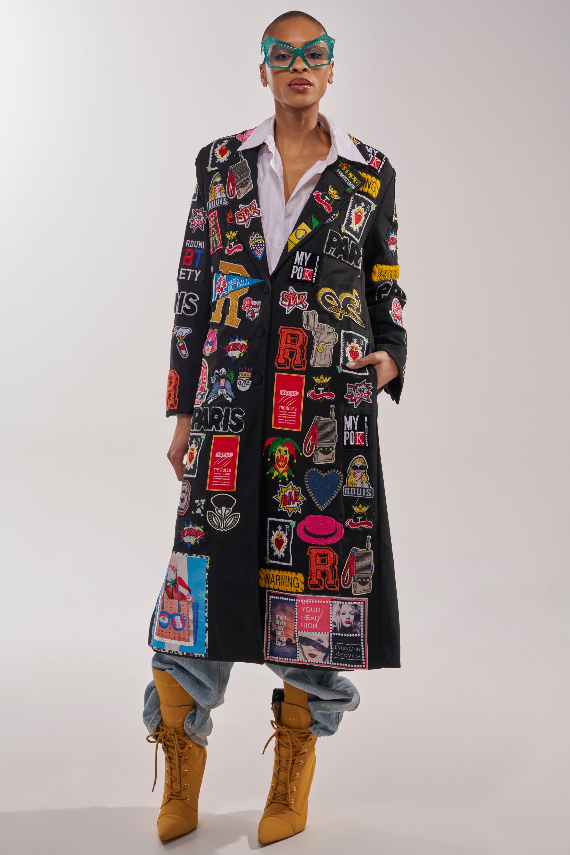 A stylish person wears the EVERYWHERE I AM PATCHWORK TRENCH, a black coat adorned with colorful patches and logos, paired with a white shirt, loose blue jeans tucked into tan lace-up boots, and blue cat-eye glasses. 4