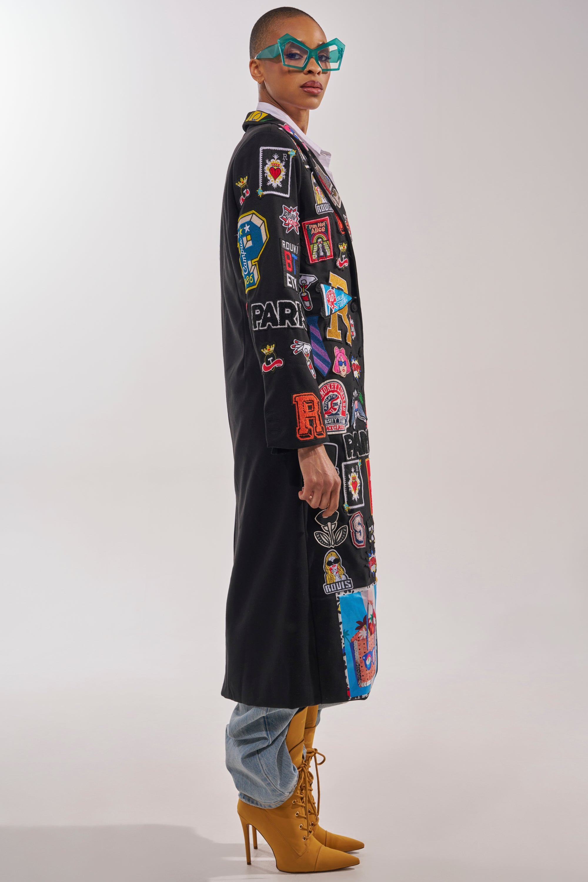 A person in the EVERYWHERE I AM PATCHWORK TRENCH, large blue glasses, light blue jeans, and tan high-heeled boots stands against a plain light background. 5
