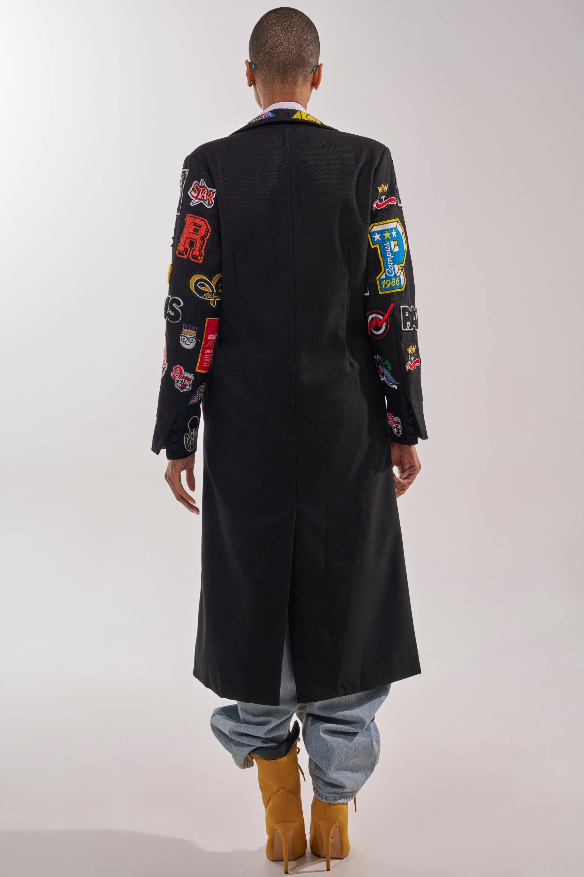 A person stands facing away in the EVERYWHERE I AM PATCHWORK TRENCH—black with colorful sleeve patches—paired with light blue jeans and tan heeled boots against a plain, light background. 7