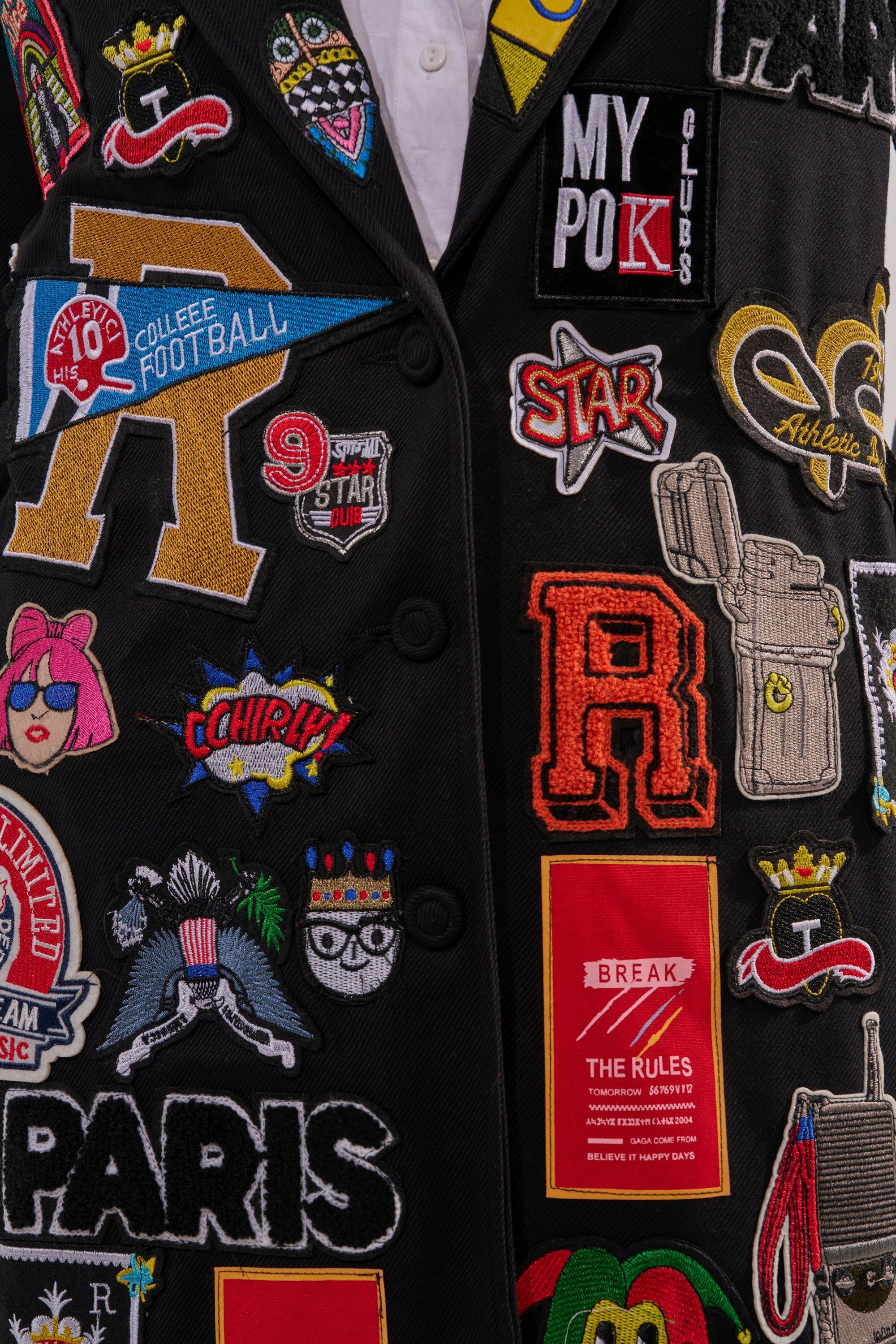 The EVERYWHERE I AM PATCHWORK TRENCH is a black trench coat adorned with colorful embroidered letters, symbols, “PARIS,” “COLLEGE FOOTBALL,” a lion, and badges—perfect statement outerwear for bold style. 8