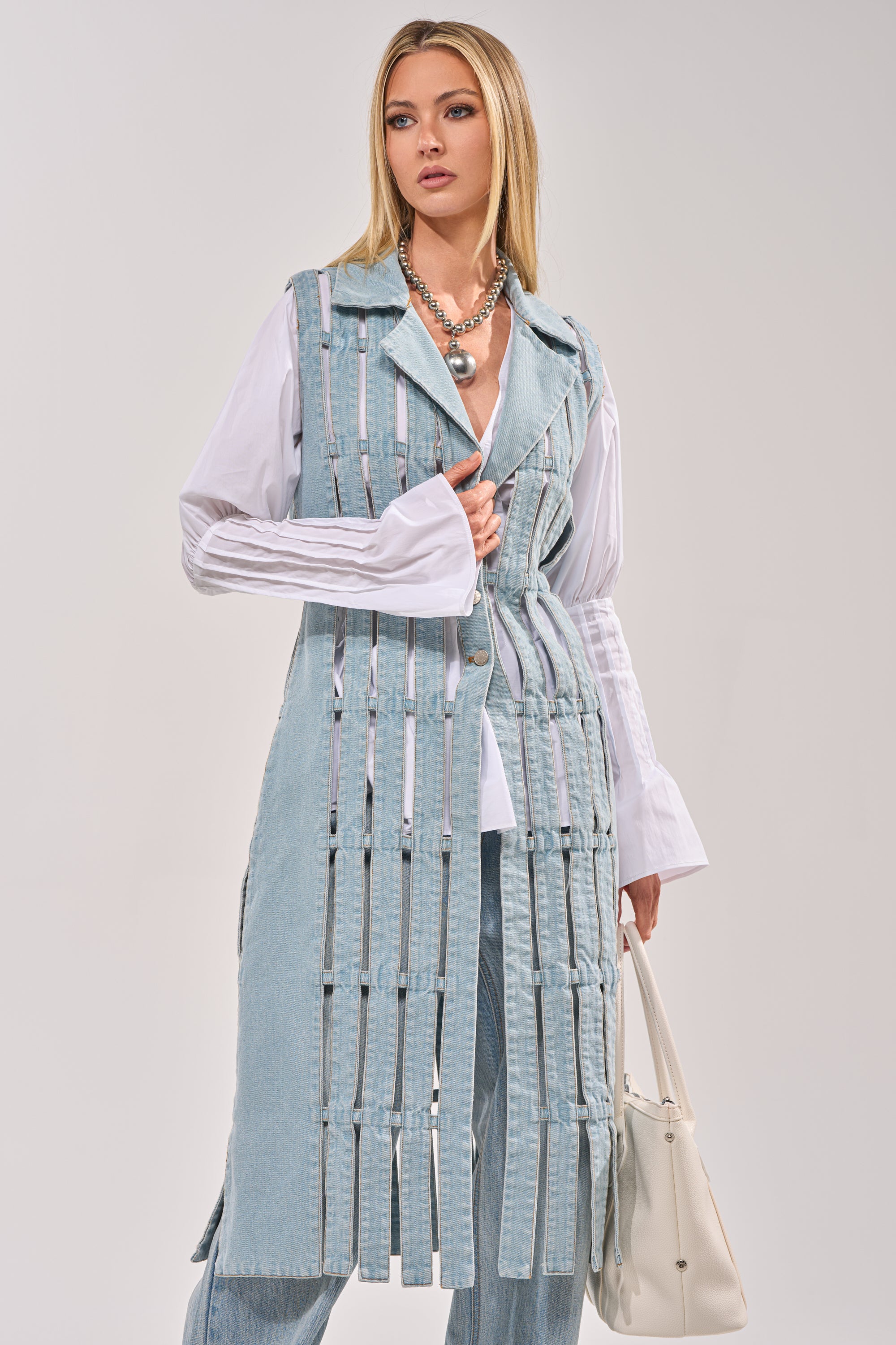 A woman showcases the RIGHT THROUGH ME CUTOUT DENIM VEST with vertical cutouts, styled over a white blouse and light blue jeans, accessorized with a white handbag and a chunky silver necklace. 0