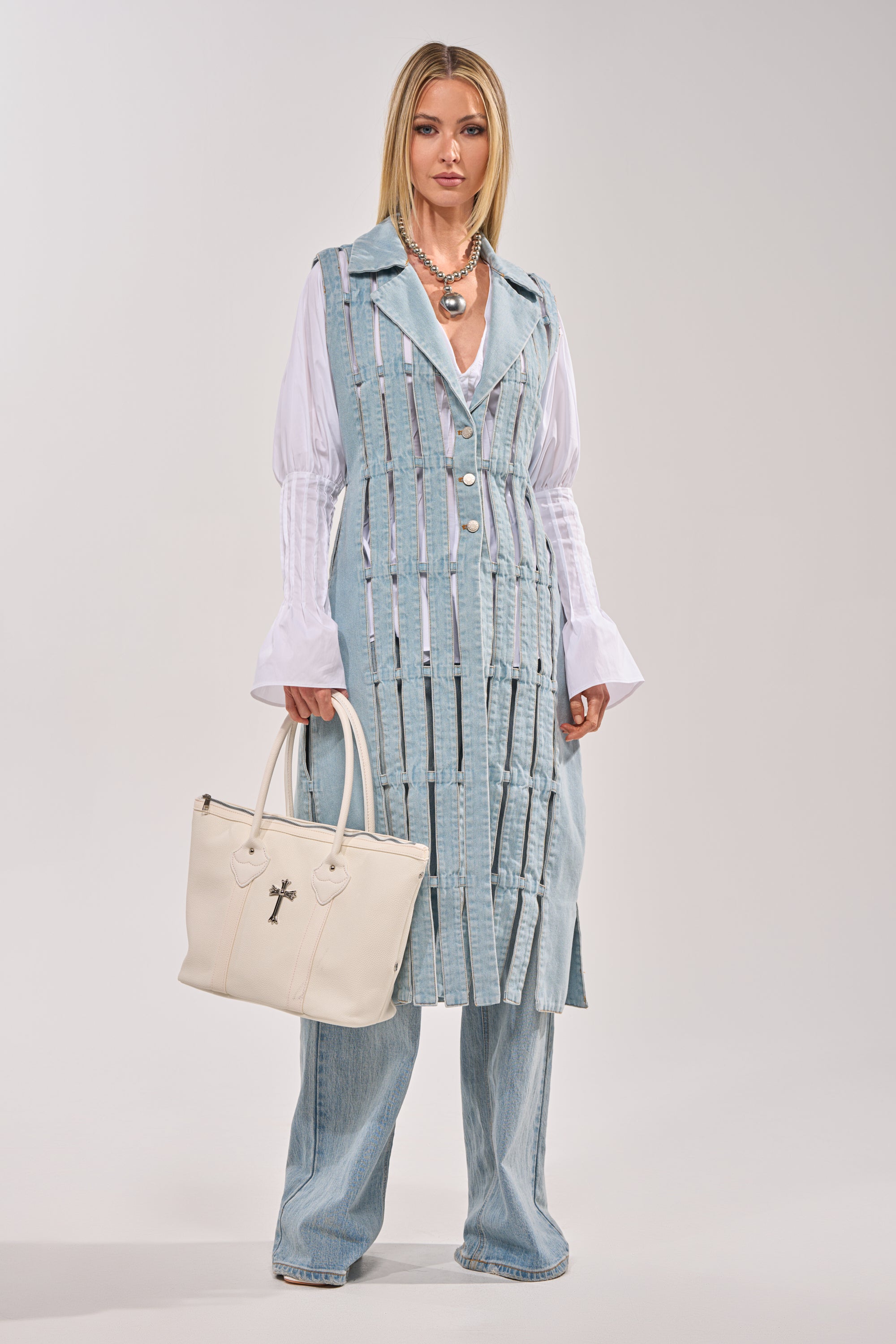 A woman stands against a plain background wearing the RIGHT THROUGH ME CUTOUT DENIM VEST, styled over a white blouse and wide-leg jeans. She accessorizes with a large white handbag and layered necklaces. 9