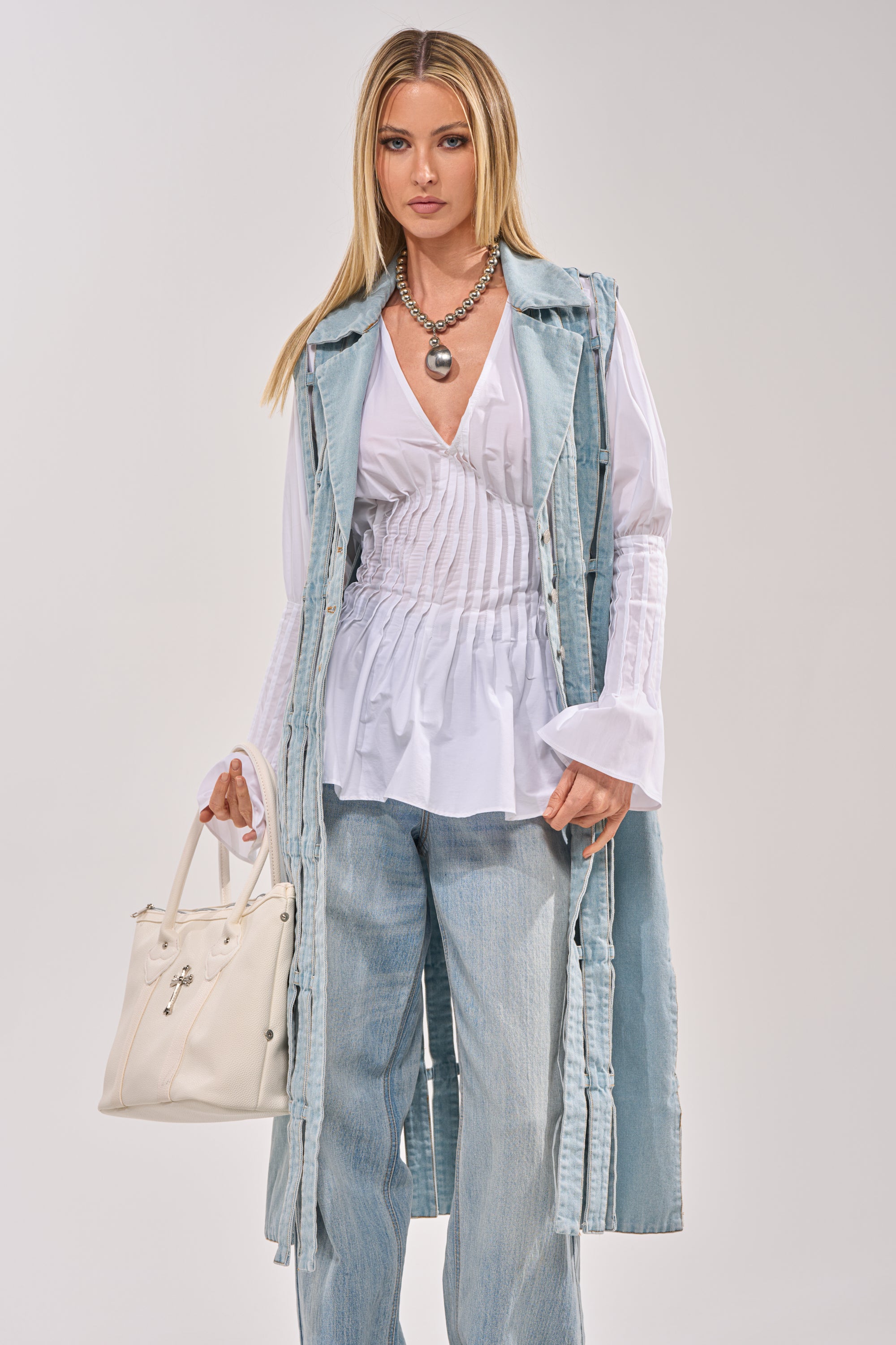 A woman with long blonde hair wears the RIGHT THROUGH ME CUTOUT DENIM VEST over a white blouse and light blue jeans, accessorized with a chunky necklace and white handbag, standing against a plain light background. 1