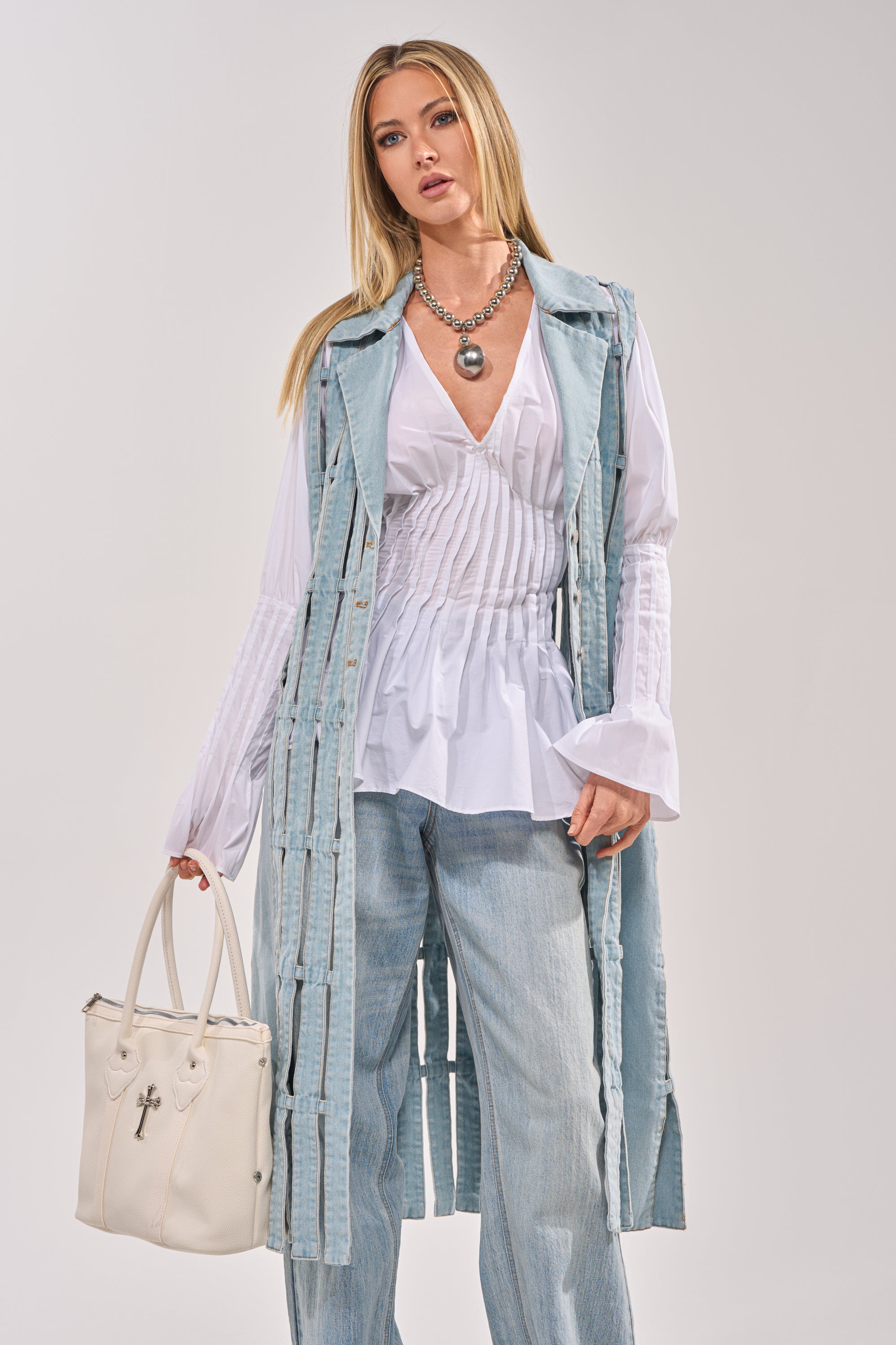 A woman with long blonde hair wears the RIGHT THROUGH ME CUTOUT DENIM VEST over a white ruched blouse and light blue jeans, accessorized with a white handbag and chunky silver necklace, posing against a light gray background. 2