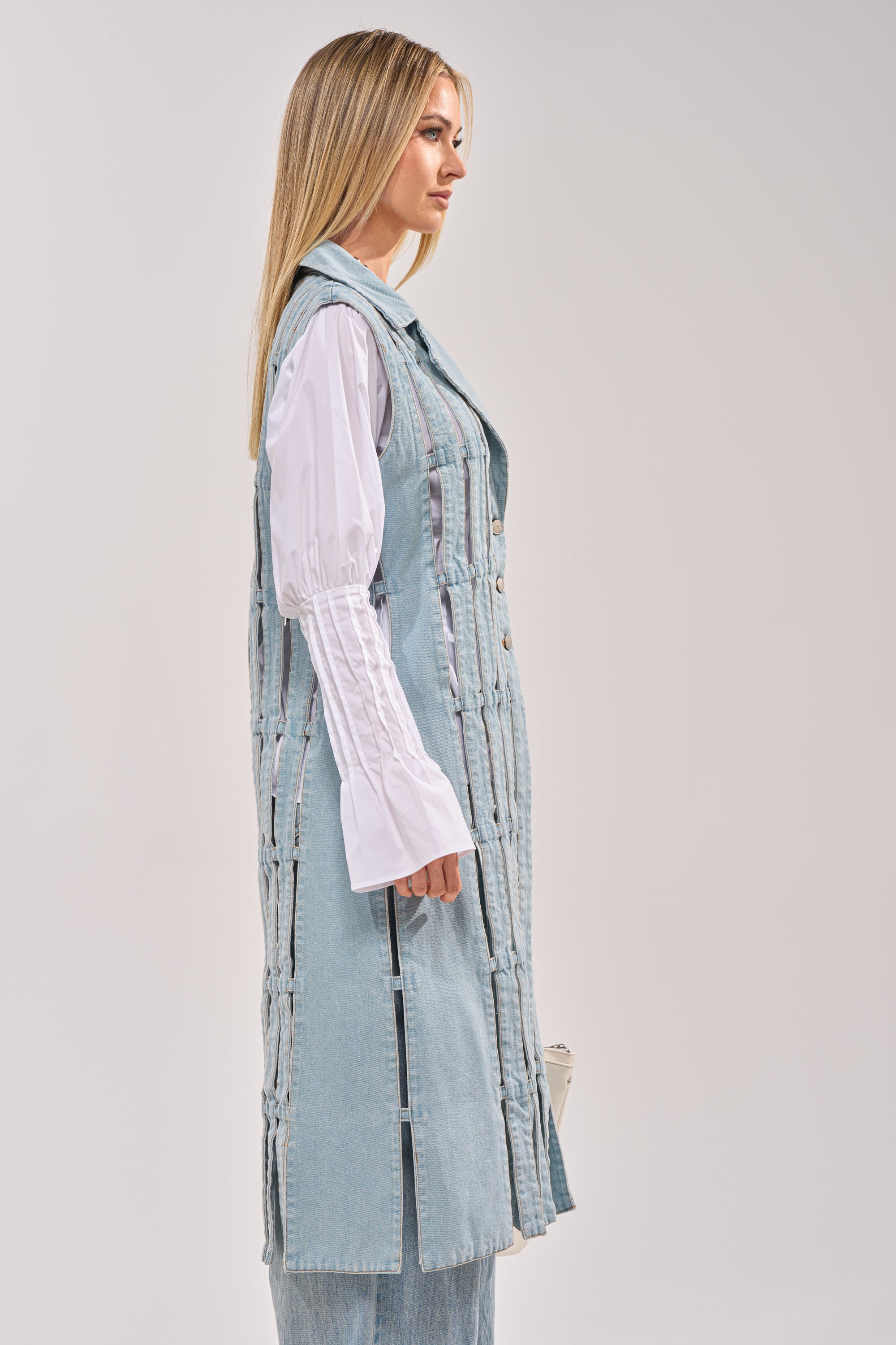 A woman with long blonde hair stands in profile, wearing the RIGHT THROUGH ME CUTOUT DENIM VEST in light blue over a white, long-sleeved shirt with flared cuffs and matching light blue pants, set against a plain background. 5