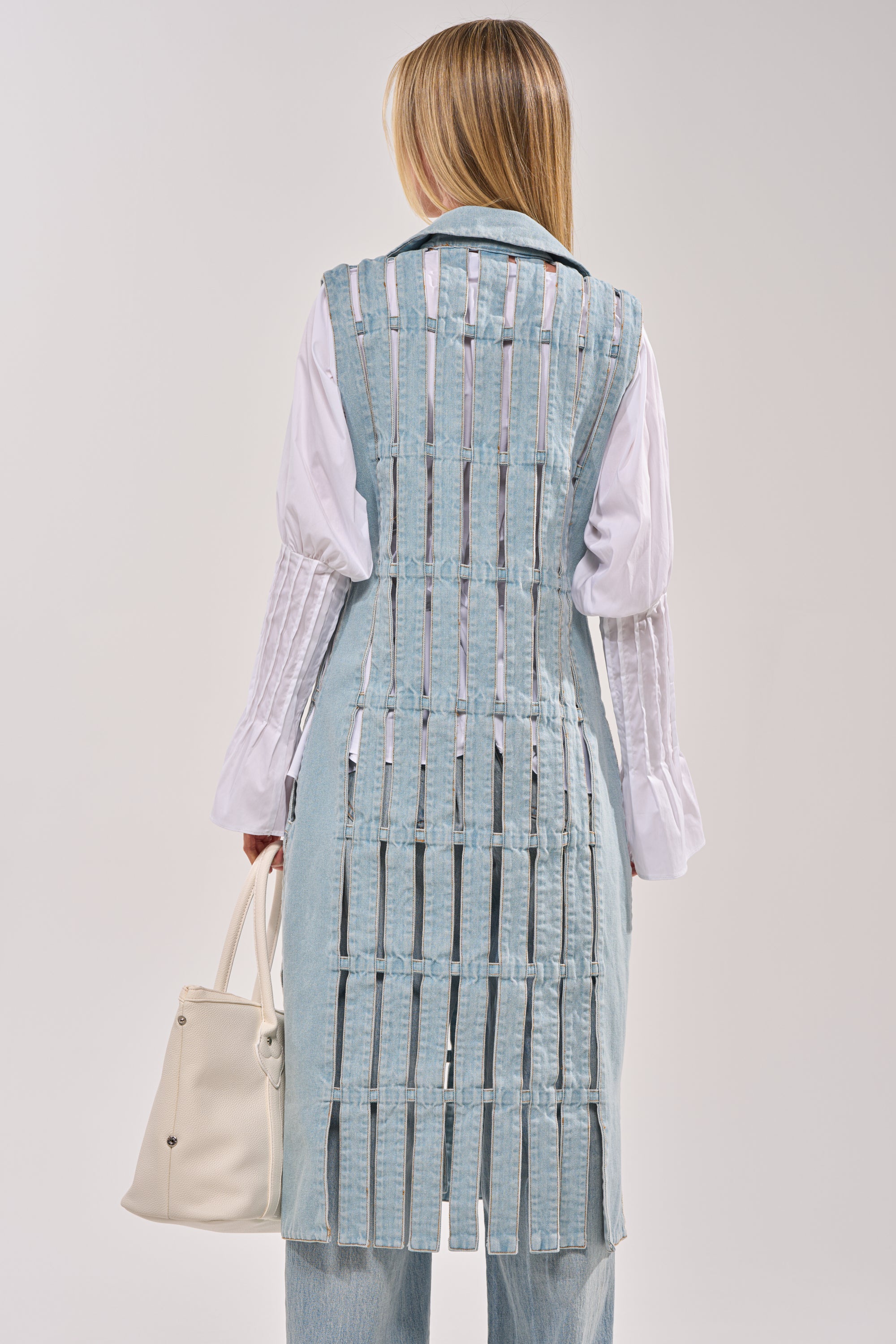 A person with long blonde hair, seen from behind, wears the RIGHT THROUGH ME CUTOUT DENIM VEST—featuring a unique open lattice back—over a white shirt and light blue jeans while holding a white bag. 6