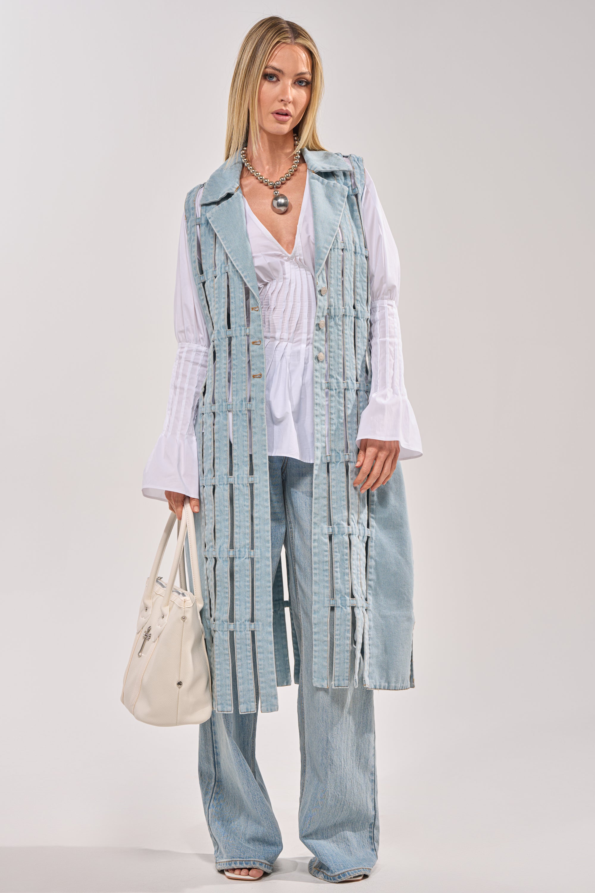 A woman stands against a light background wearing the RIGHT THROUGH ME CUTOUT DENIM VEST styled over a white flared-sleeve blouse, wide-leg jeans, layered necklaces, and holding a white handbag. 7