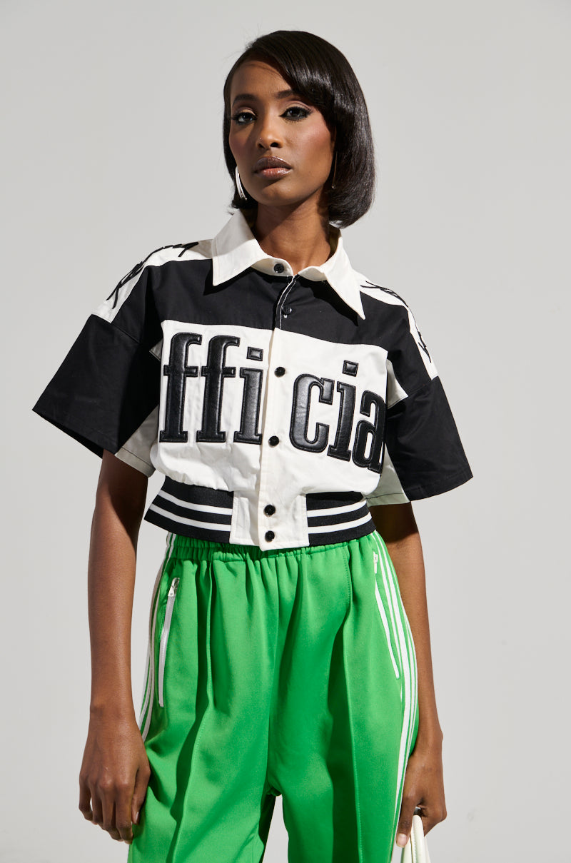 A person wearing the Official Duty Cropped Button Down, featuring a color block pattern with "official" emblazoned on it, and bright green pants with an elasticized waistband stands against a neutral background. They have short black hair and look confidently at the camera.
