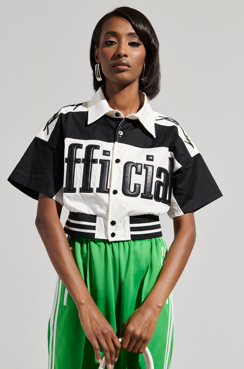 A person stands confidently in the OFFICIAL DUTY CROPPED BUTTON DOWN, featuring a bold text design in black and white, paired with vibrant green pants that have an elasticized waistband. The plain background highlights the stylish, color-block pattern of the outfit.