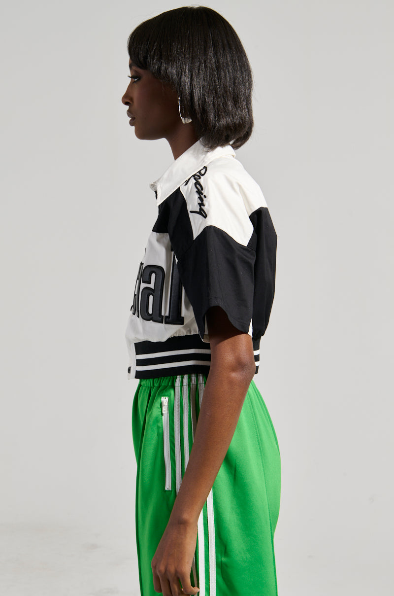 A person with short black hair is wearing the OFFICIAL DUTY CROPPED BUTTON DOWN, a black and white color block shirt with writing on the shoulder, paired with bright green striped pants featuring an elasticized waistband. They stand in profile against a plain gray background.