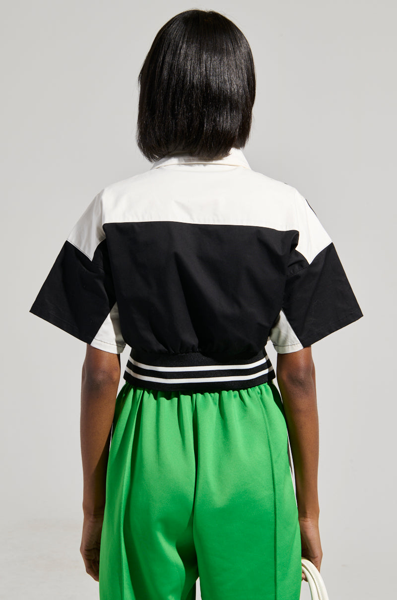 A person with short, dark hair stands with their back to the camera, showcasing a stylish OFFICIAL DUTY CROPPED BUTTON DOWN top in a color block pattern, paired with vibrant green pants featuring an elasticized waistband against a neutral gray background.
