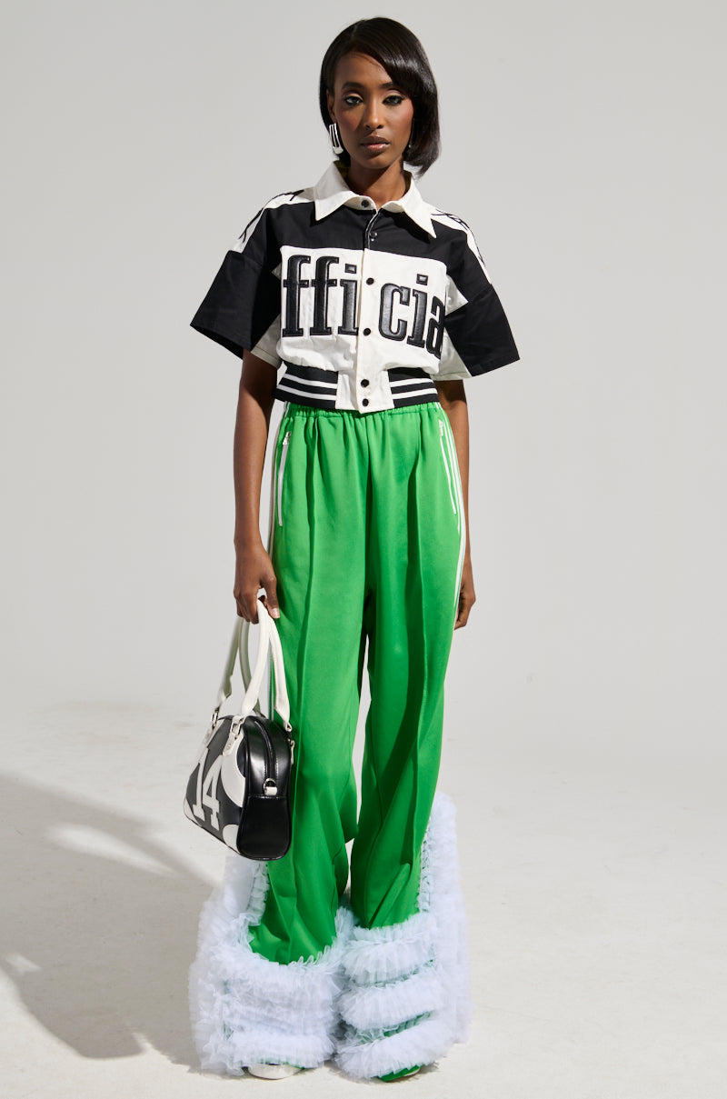 A person models an outfit featuring the "OFFICIAL DUTY CROPPED BUTTON DOWN" in black and white, with the word "offici" partially visible. It is paired with bright green pants that have fluffy white trim at the hem, and the ensemble is completed with a chic black and white handbag.
