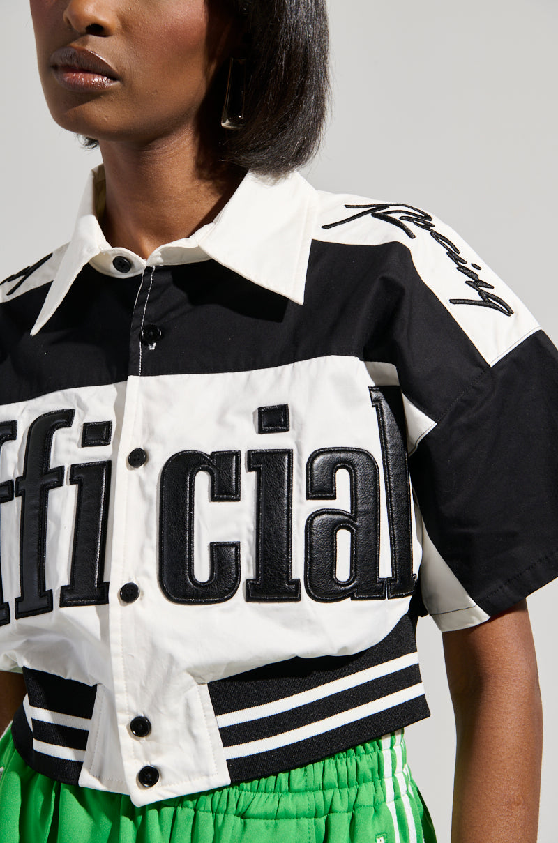 A person wears the OFFICIAL DUTY CROPPED BUTTON DOWN, showcasing a black and white color block pattern with "official" partially visible in bold letters. Their shoulder-length hair pairs well with green pants featuring white stripes and an elasticized waistband for comfort.
