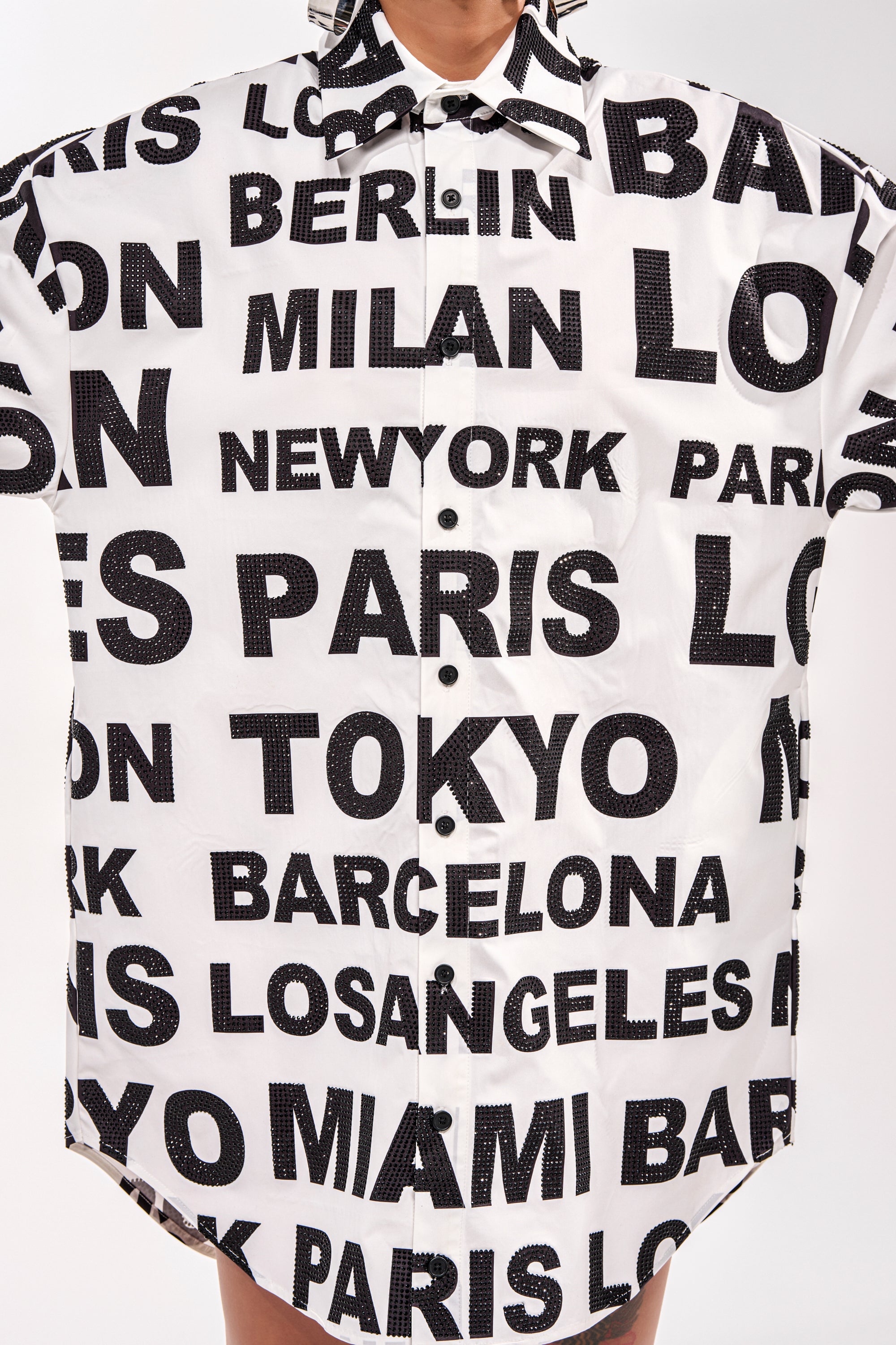 A person wears the TIME AND PLACE POPLIN MINI DRESS, a black and white mini shirt dress with bold city name text including Paris, Tokyo, Berlin, Milan, and New York. Only the torso and part of the neck are visible. 10