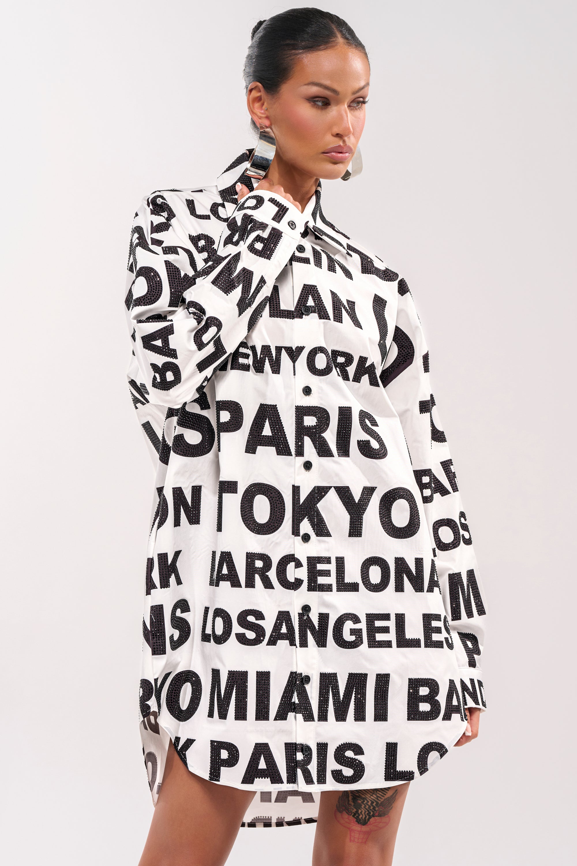 A woman wears the TIME AND PLACE POPLIN MINI DRESS—a black and white graphic mini shirt dress featuring large city names—while standing against a plain, light background. 2