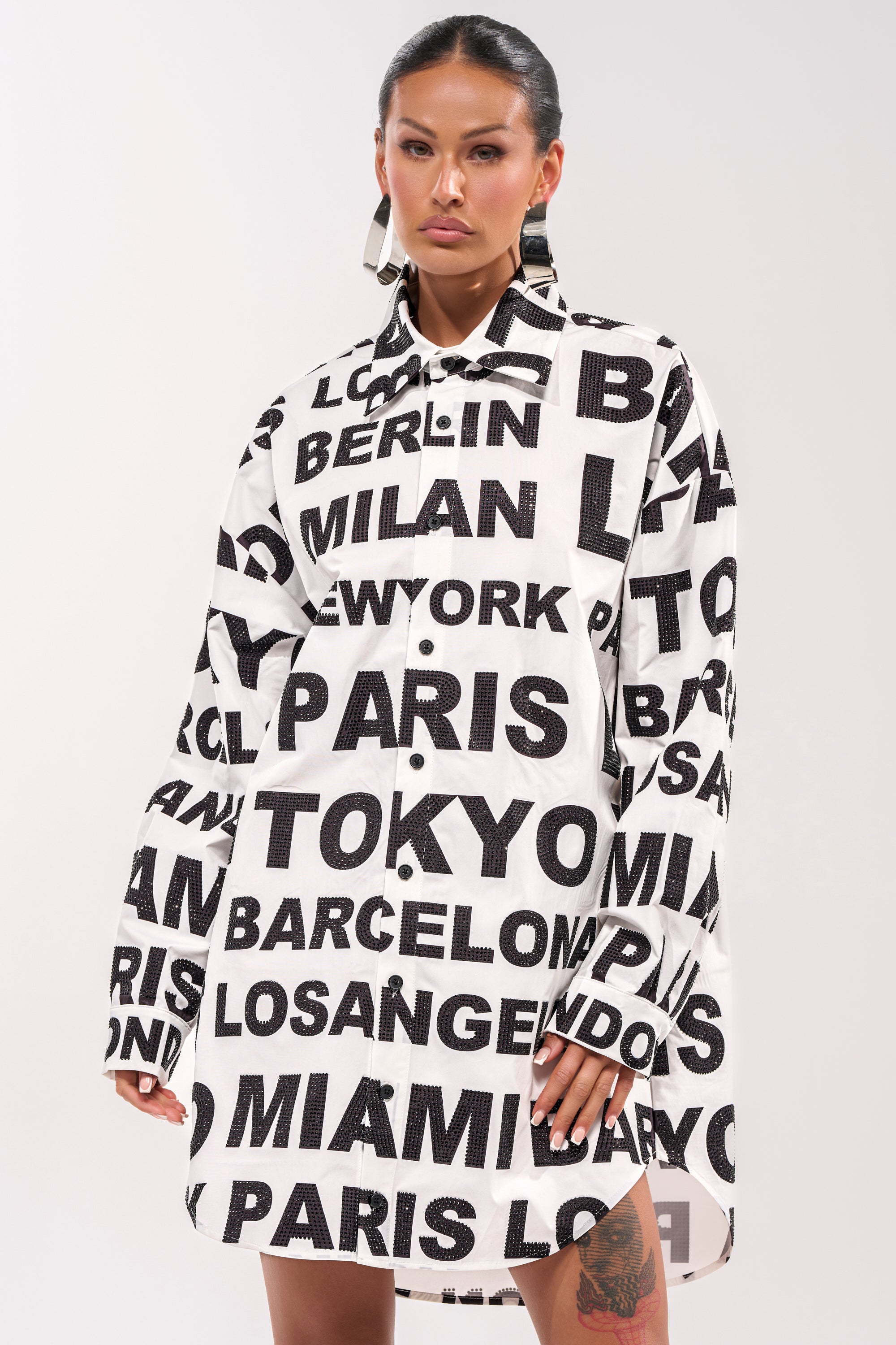 A person wears the TIME AND PLACE POPLIN MINI DRESS, a black and white oversized shirt dress with large city names like Tokyo, Paris, Milan, Miami, and New York printed on it. The background is plain and light-colored. 3