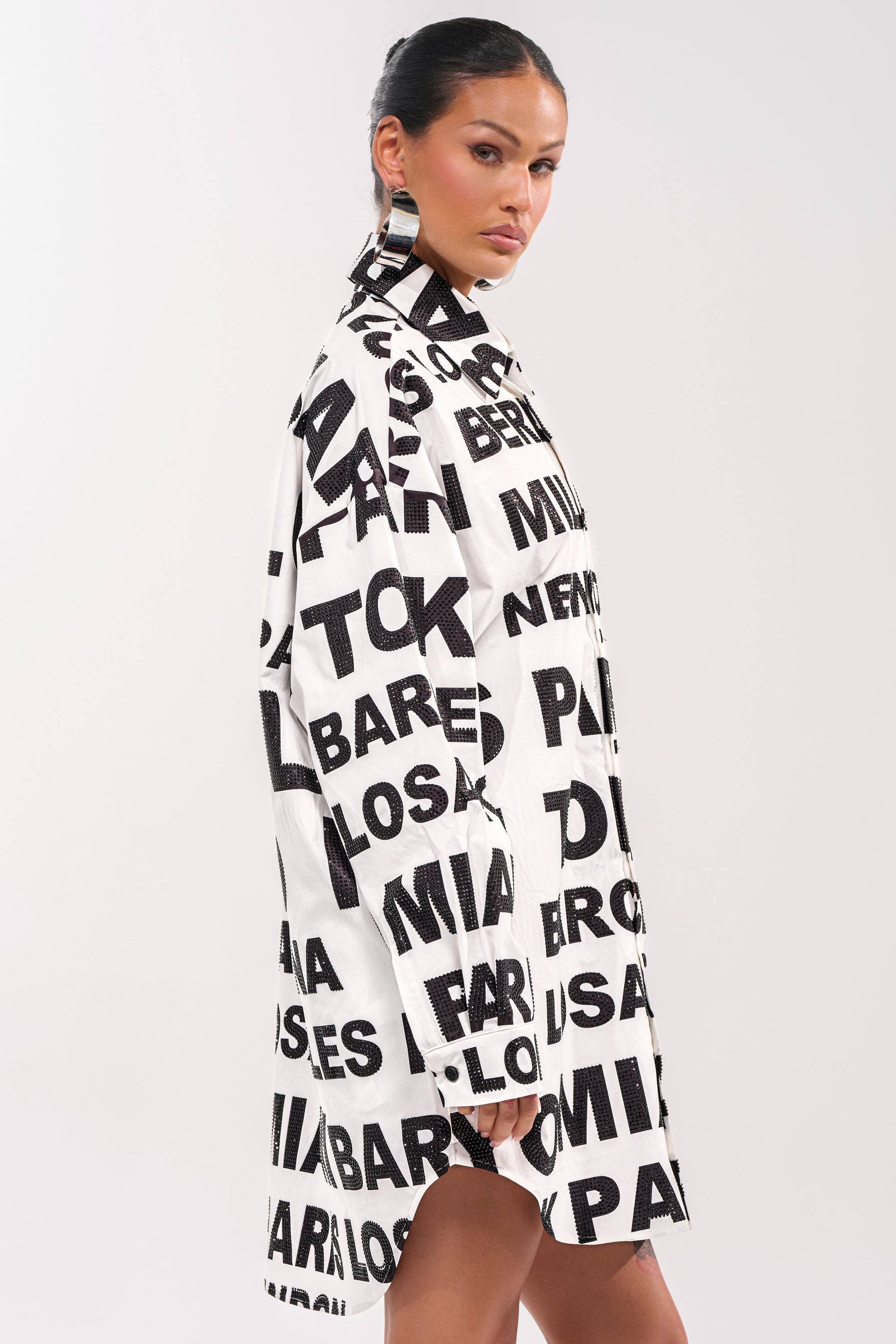 A woman wearing the TIME AND PLACE POPLIN MINI DRESS in a bold black and white graphic print stands sideways against a plain background, looking confidently at the camera with slicked-back hair and large geometric earrings. 4