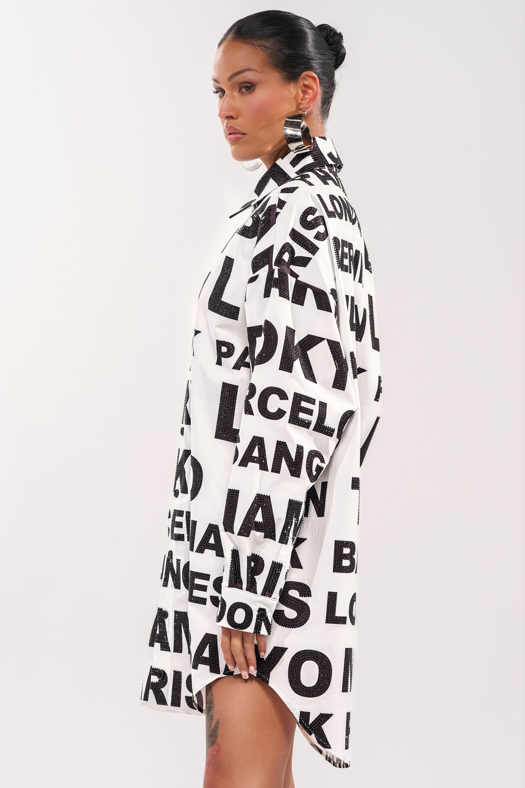 A woman with slicked-back hair and bold hoop earrings models the TIME AND PLACE POPLIN MINI DRESS, featuring a black and white graphic print of letters and city names. She faces sideways against a plain light background. 5