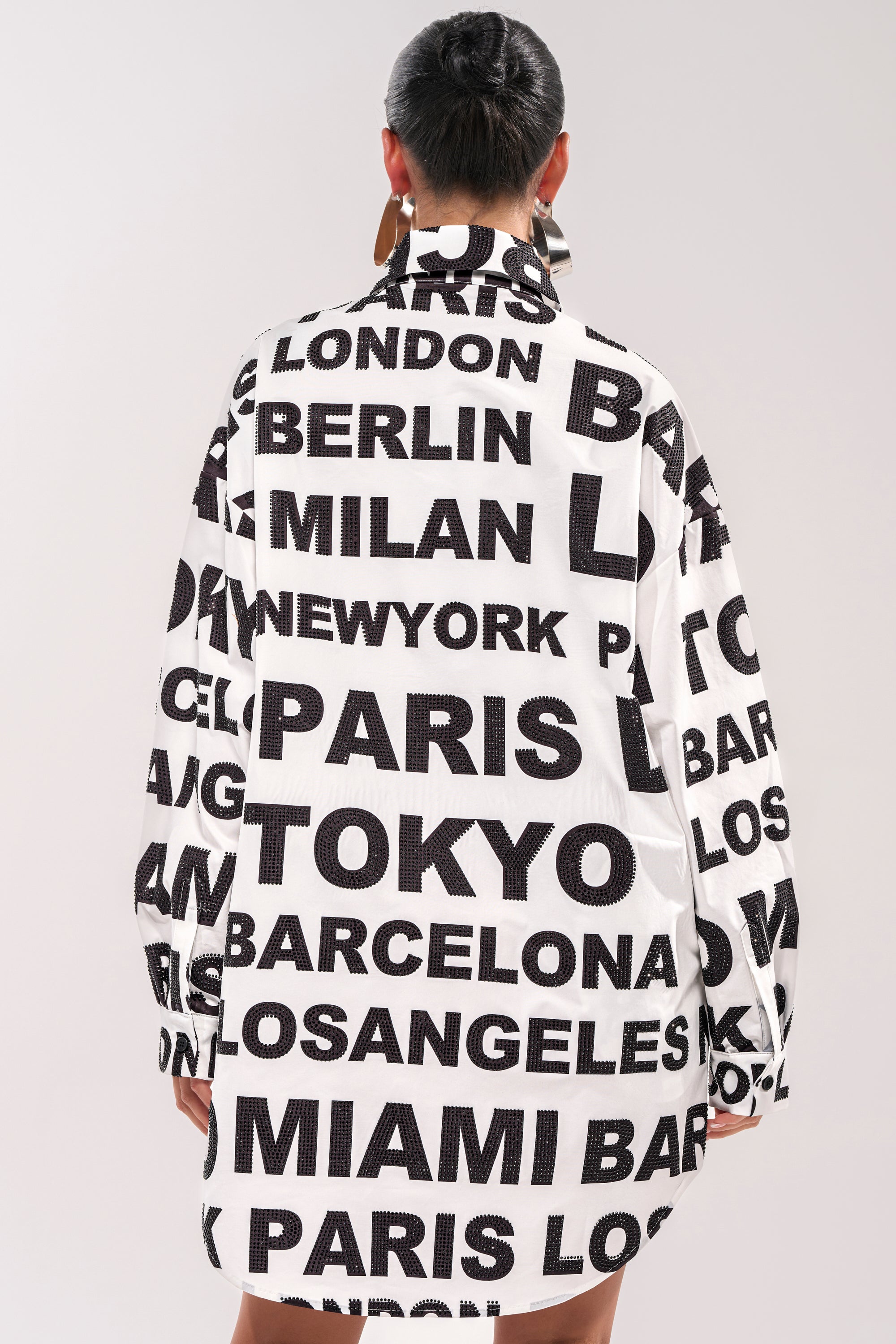 A person with short dark hair, seen from behind, wears the TIME AND PLACE POPLIN MINI DRESS—a black and white graphic mini dress with long sleeves and bold city names like Paris, Tokyo, London, and New York. 6