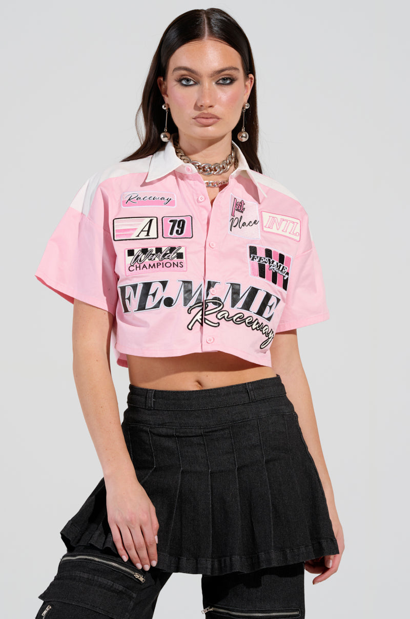 A person with long dark hair sports a button-down pink poplin shirt from the "Femme Racing" collection, accentuated with racing-themed details. They pair it elegantly with a black pleated skirt against a plain light gray backdrop.