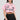 A person with long dark hair sports a button-down pink poplin shirt from the "Femme Racing" collection, accentuated with racing-themed details. They pair it elegantly with a black pleated skirt against a plain light gray backdrop.