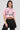 A person with long dark hair sports a button-down pink poplin shirt from the "Femme Racing" collection, accentuated with racing-themed details. They pair it elegantly with a black pleated skirt against a plain light gray backdrop.