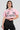 The woman is dressed in the FEMME RACING BUTTON DOWN POPLIN SHIRT IN PINK, a cotton poplin button-up crop shirt featuring embroidered patchwork and racing-themed patches. She pairs it with a black pleated skirt and accessorizes with hoop earrings and a chain choker against a simple gray backdrop.