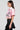 A woman with long hair is wearing the Femme Racing Button Down Poplin Shirt in pink, paired with a black pleated skirt. She stands in profile against a plain background.