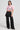 A woman wearing the Femme Racing button-down poplin shirt in pink, featuring graphic designs and an embroidered patchwork. She pairs it with a black pleated mini skirt, black cargo pants with zippers, and chunky black shoes. Positioned against a plain background, she showcases a bold, stylish fashion look.