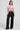 A woman stands wearing the Femme Racing Button Down Poplin Shirt in pink, paired with black wide-leg cargo pants featuring multiple pockets. She complements her outfit with black platform shoes and has long brown hair. The plain white backdrop accentuates her unique style.