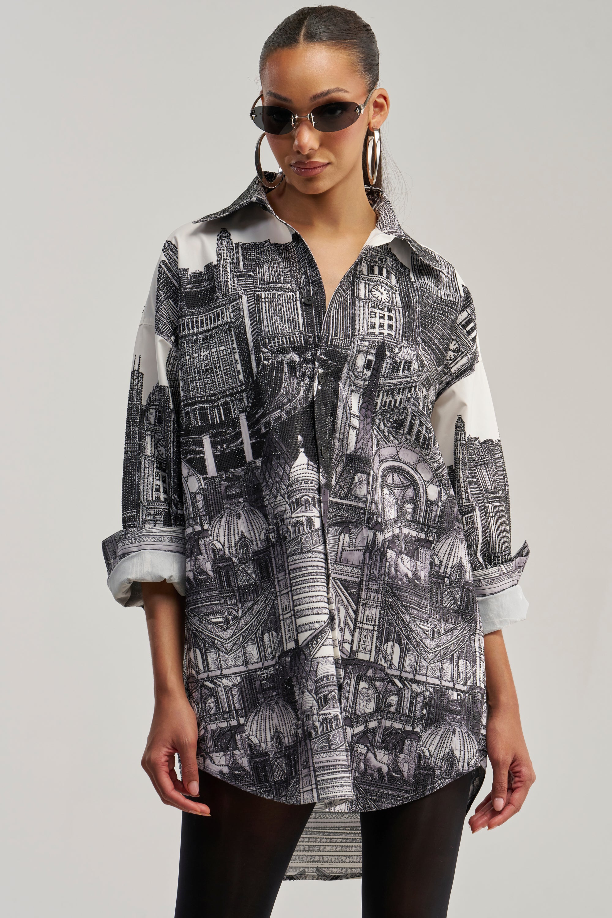 Wearing sunglasses and large hoop earrings, a woman models the ALL AROUND THE WORLD POPLIN MINI DRESS, a graphic shirt dress adorned with detailed black and white architectural prints, standing against a plain background.
