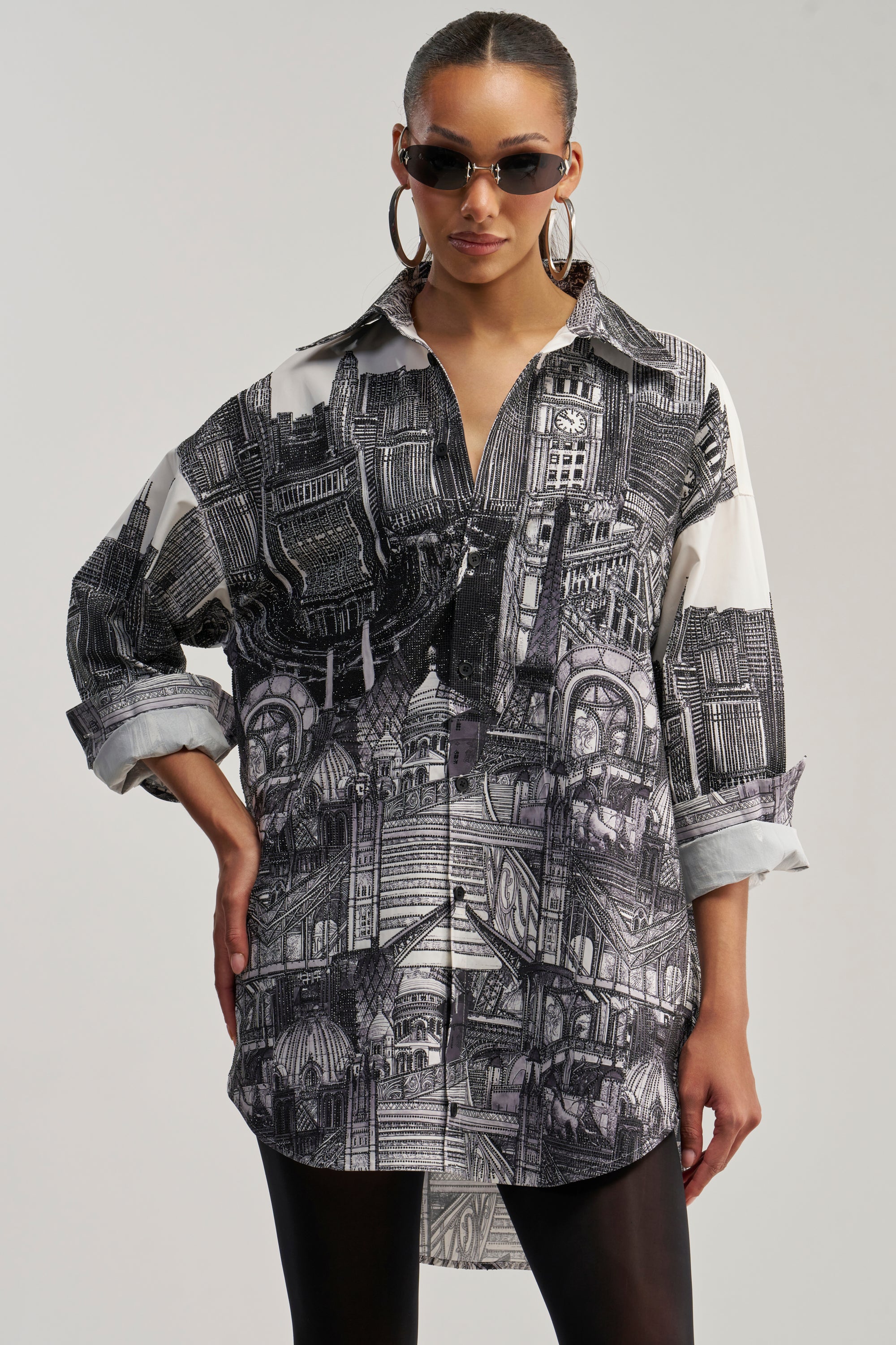 A woman wearing sunglasses and hoop earrings confidently models the ALL AROUND THE WORLD POPLIN MINI DRESS—a black and white mini dress featuring intricate architectural patterns and rolled-up sleeves—against a plain background.