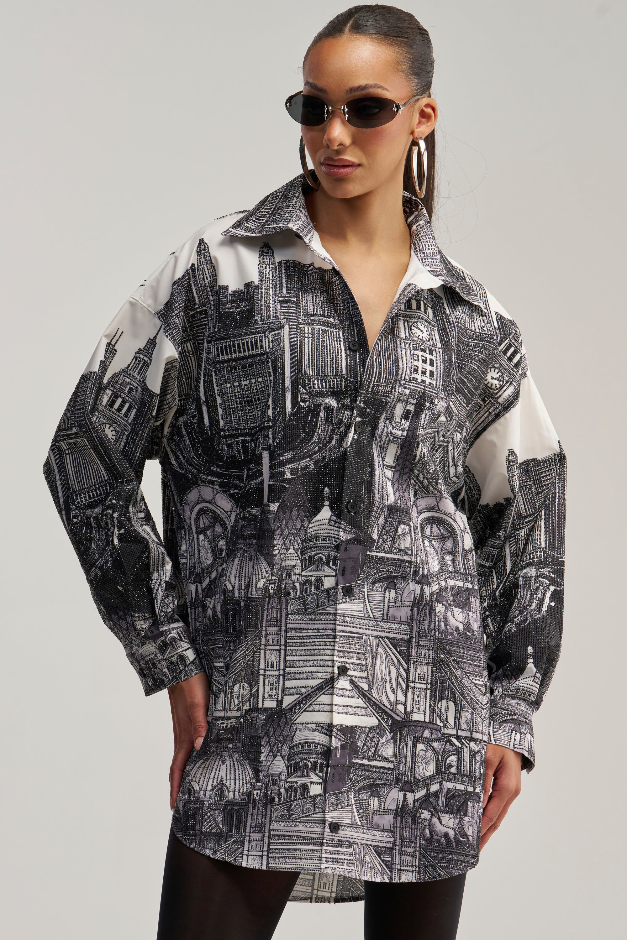 A woman stands with one hand on her hip against a neutral background, wearing sunglasses, large hoop earrings, and the ALL AROUND THE WORLD POPLIN MINI DRESS—a black-and-white graphic shirt dress featuring architectural city illustrations.