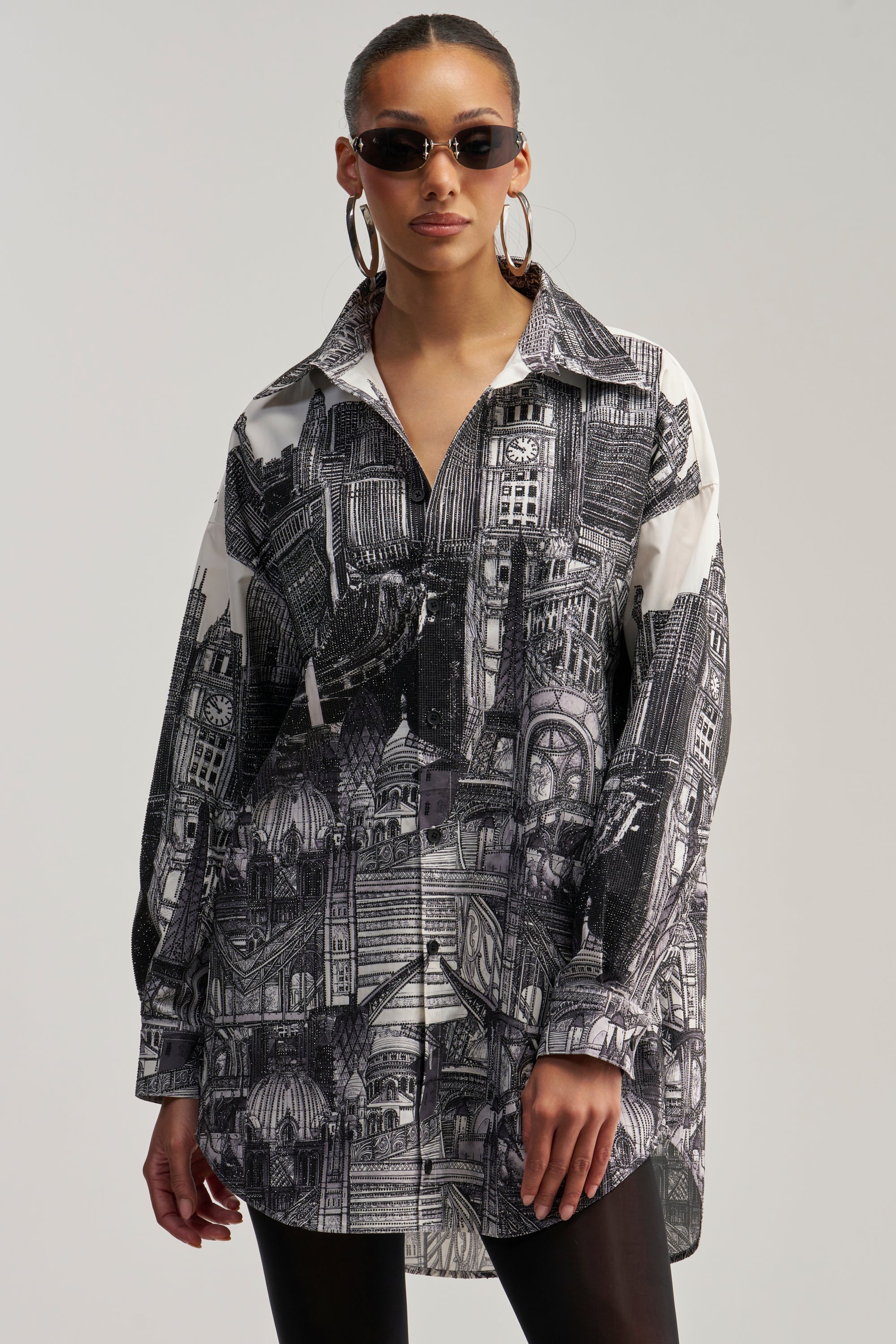A woman wearing round sunglasses, large hoop earrings, and the ALL AROUND THE WORLD POPLIN MINI DRESS featuring a bold black and white architectural print, stands against a plain background.