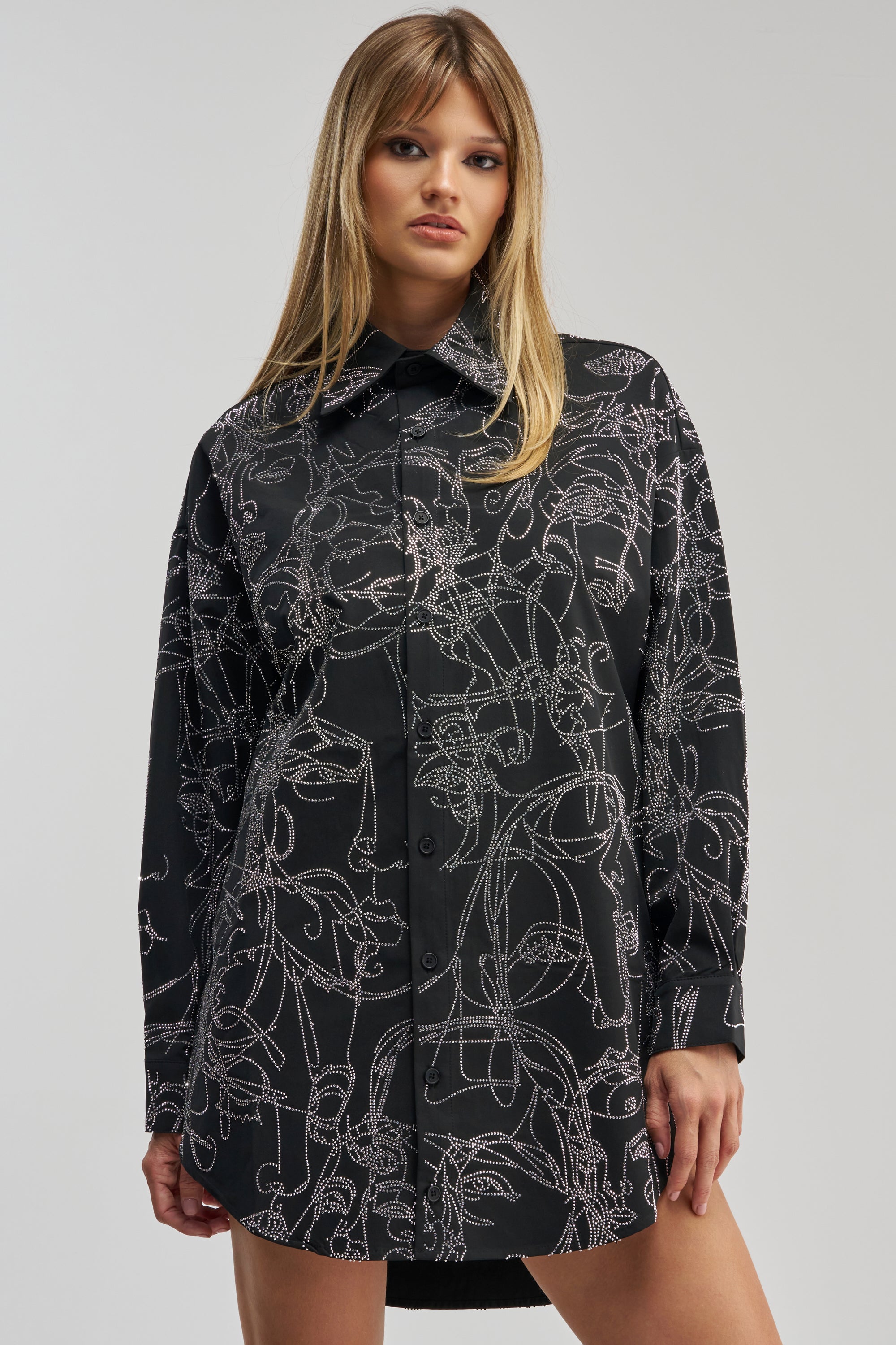 A woman faces forward, wearing the ZIG AND ZAG POPLIN MINI DRESS—a long-sleeved black mini dress with a white abstract line pattern and button-up front. Her straight blonde hair frames a neutral expression as she stands against a plain background.