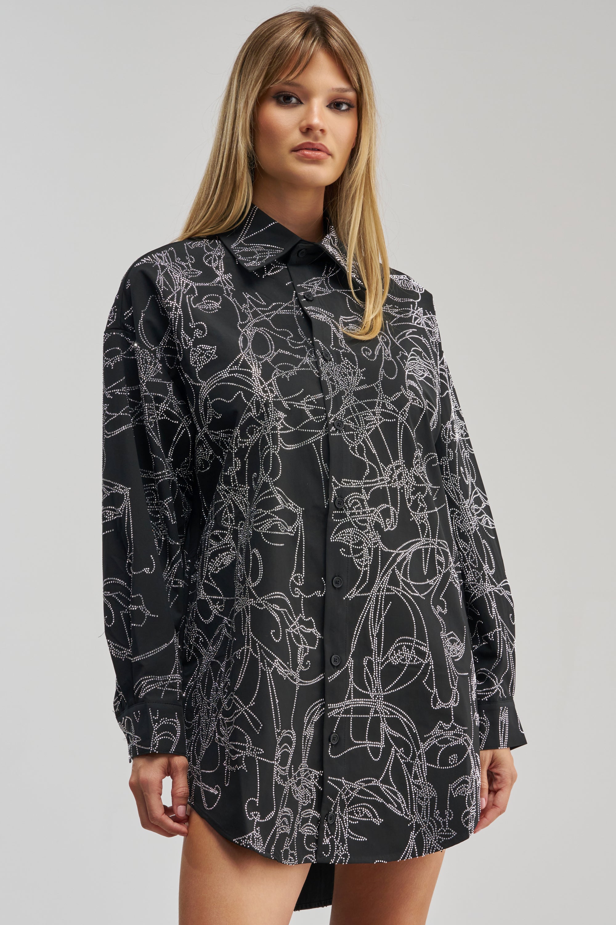 A woman with straight blonde hair and a neutral expression poses against a plain, light background wearing the ZIG AND ZAG POPLIN MINI DRESS— a long-sleeved black poplin mini dress adorned with an abstract white line art face pattern.