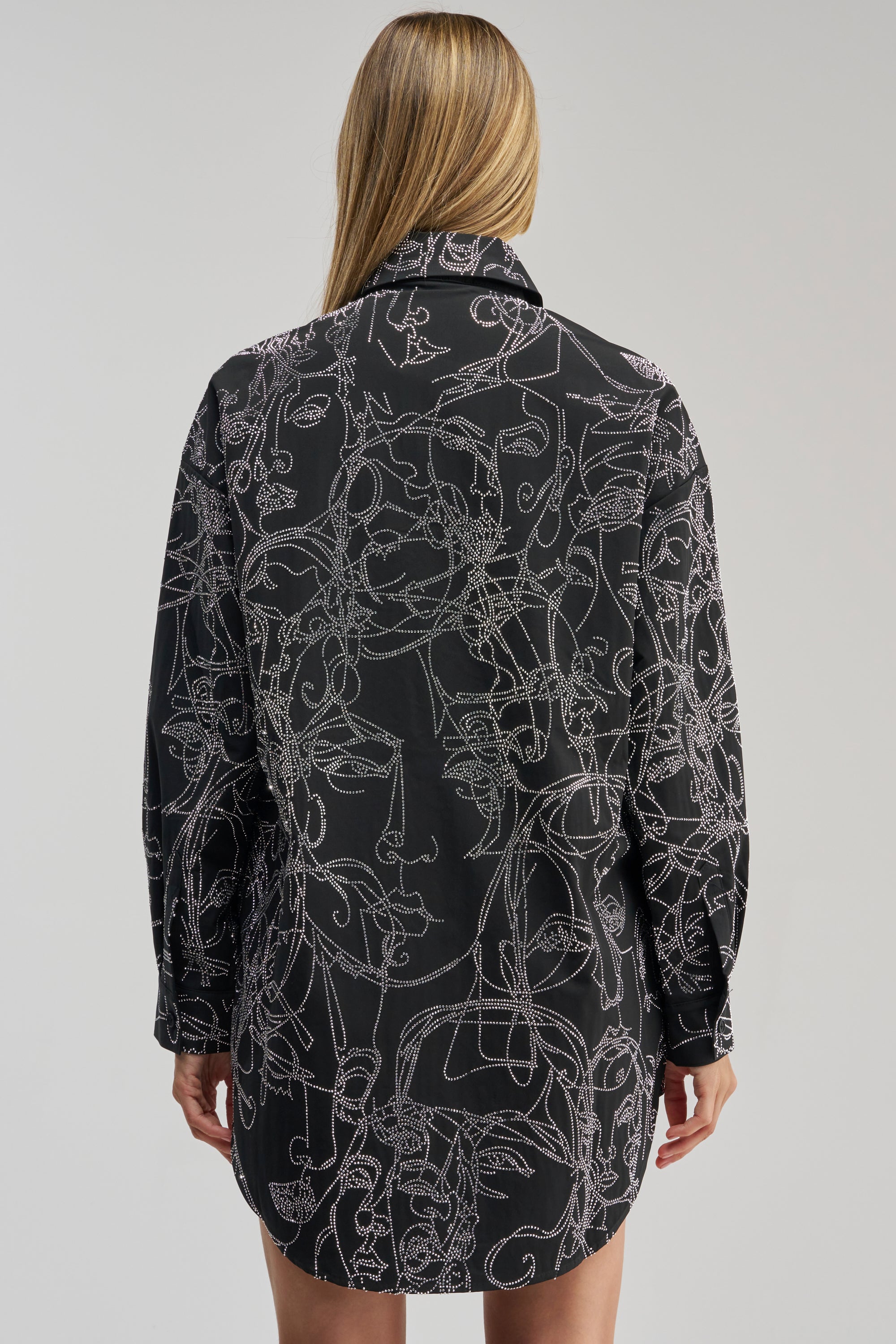 Wearing the ZIG AND ZAG POPLIN MINI DRESS, a person with long blonde hair is shown from the back against a plain, light background. The black poplin mini dress features an abstract white line art face pattern.