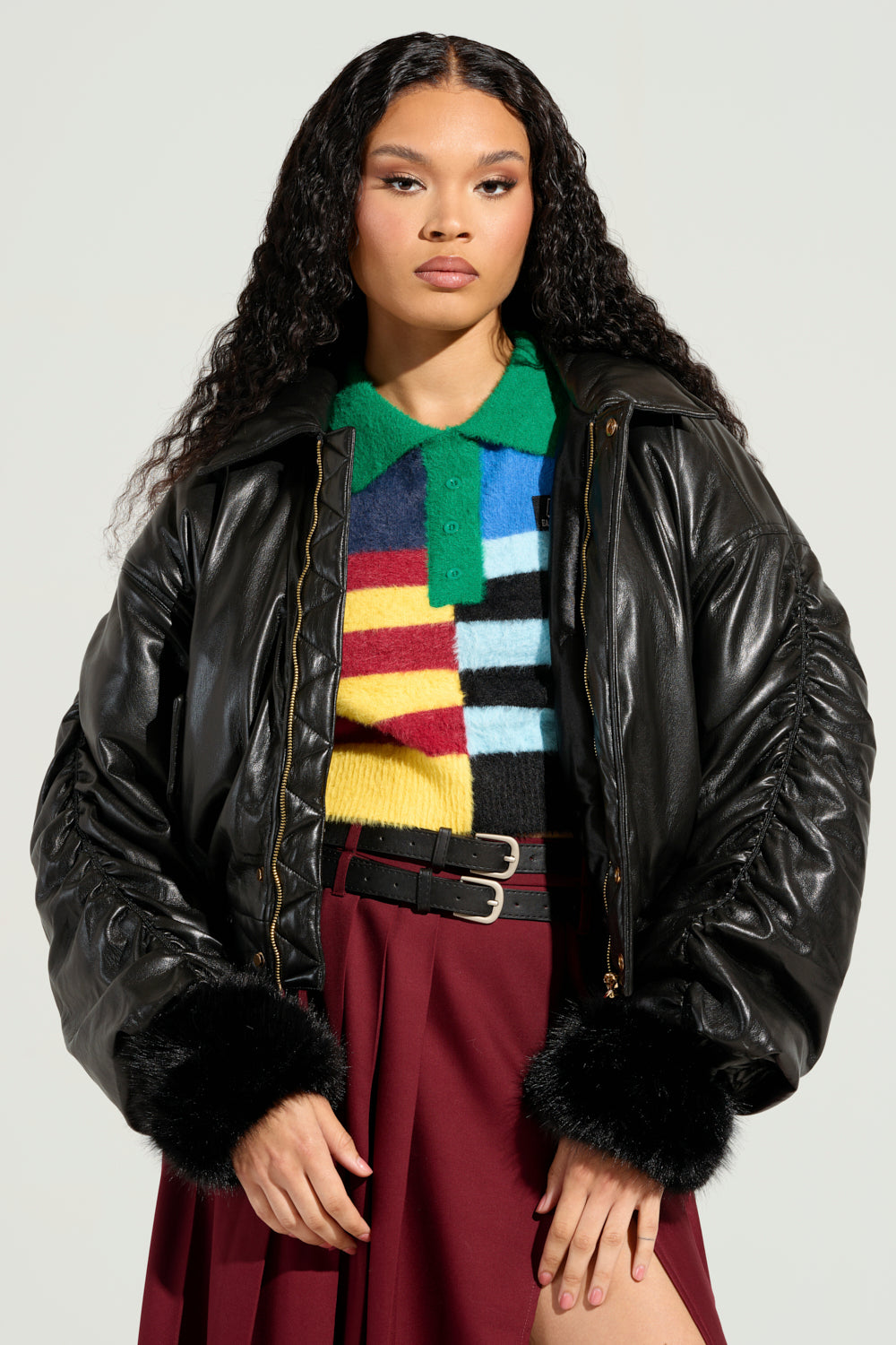 A person with long curly hair models the TOKYO DRIFTING FAUX LEATHER BOMBER, which features fur cuffs, over a colorful striped sweater. They complement this look with a burgundy skirt and exude confidence against a plain background.