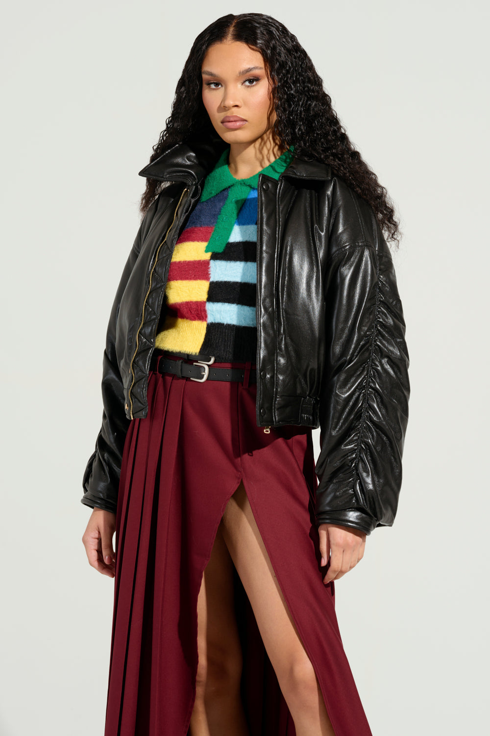 A person with long curly hair wears a TOKYO DRIFTING FAUX LEATHER BOMBER over a colorful striped sweater and a red skirt with high slits. The background is plain and light-colored, enhancing the Tokyo drifting vibe.