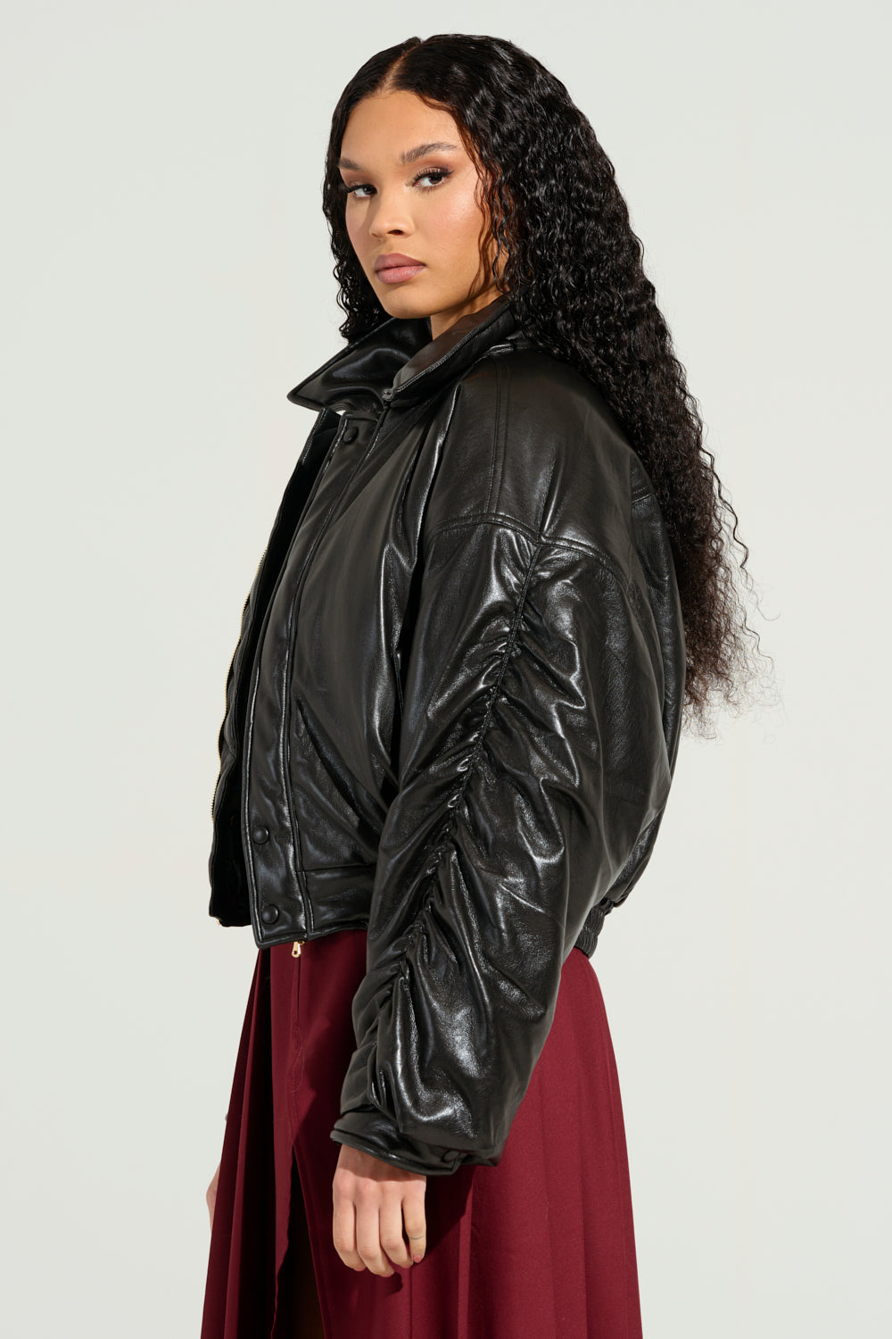 A person with long, curly hair is wearing the TOKYO DRIFTING FAUX LEATHER BOMBER and a burgundy skirt. They are standing against a plain, light background, looking to the side with a neutral expression.