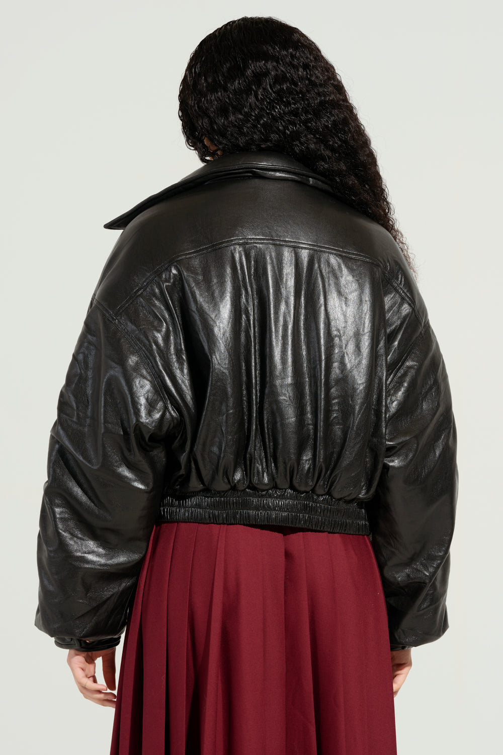 Photographed from behind, a person with long, curly hair is wearing the stylish TOKYO DRIFTING FAUX LEATHER BOMBER known for its cropped boxy silhouette and elastic waistband, paired with a burgundy skirt. The background is plain and light-colored.