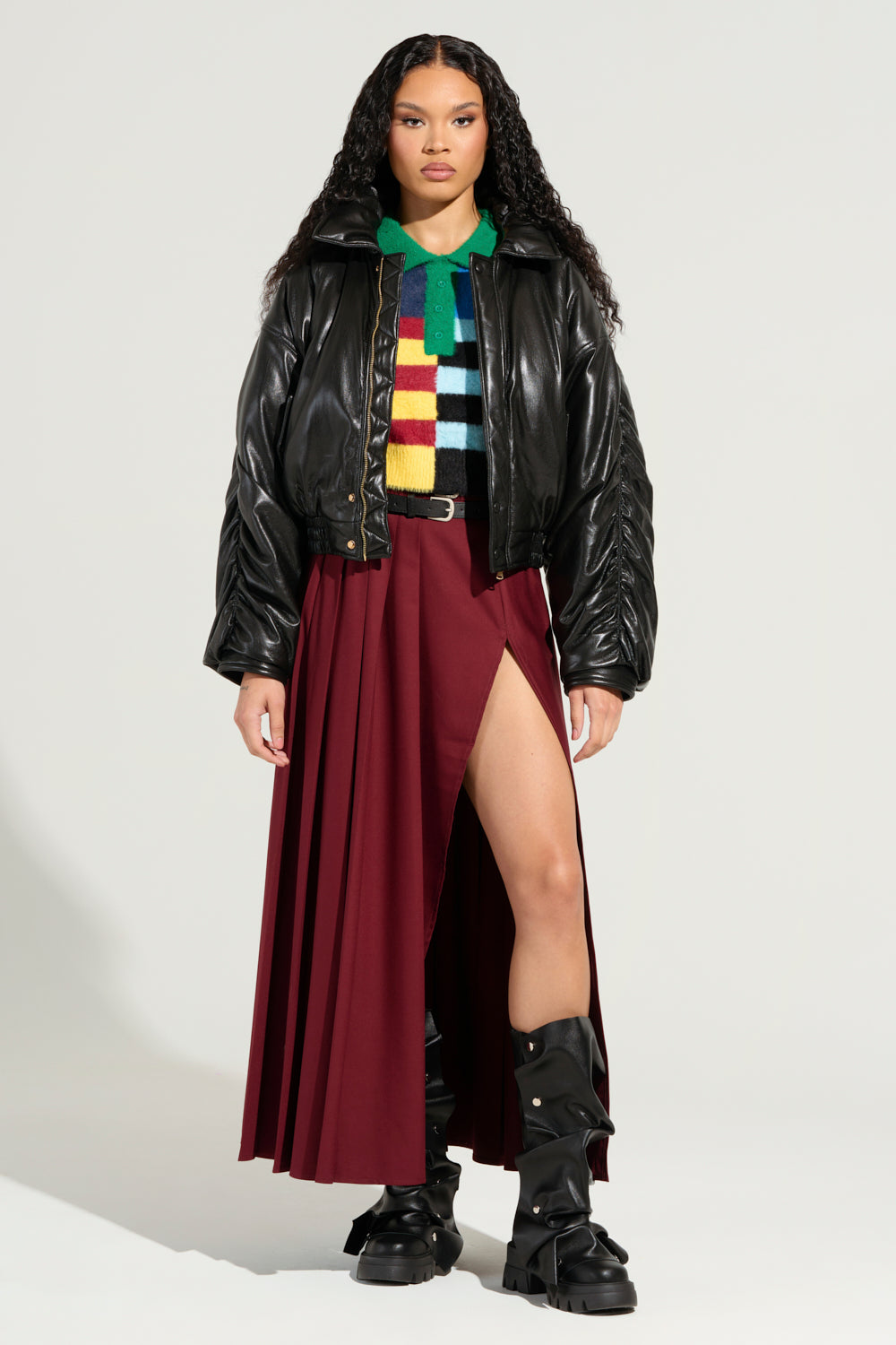 Wearing the TOKYO DRIFTING FAUX LEATHER BOMBER in black over a colorful striped sweater, paired with a burgundy skirt featuring a high slit and black boots, this ensemble captures an essence of Tokyo drifting cool. The cropped boxy silhouette enhances the confident pose against the plain light background.